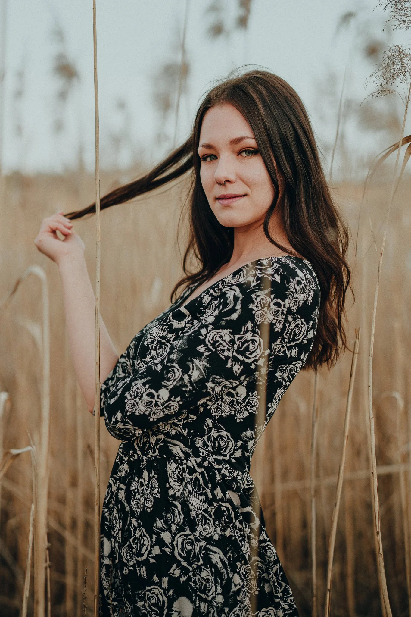 Junge Frau mit braunen Haaren in einem schwarzen Kleid mit weißen Rosen- und Totenkopf-Print, steht in einem Feld mit trockenen Gräsern, bei Sonnenuntergang.