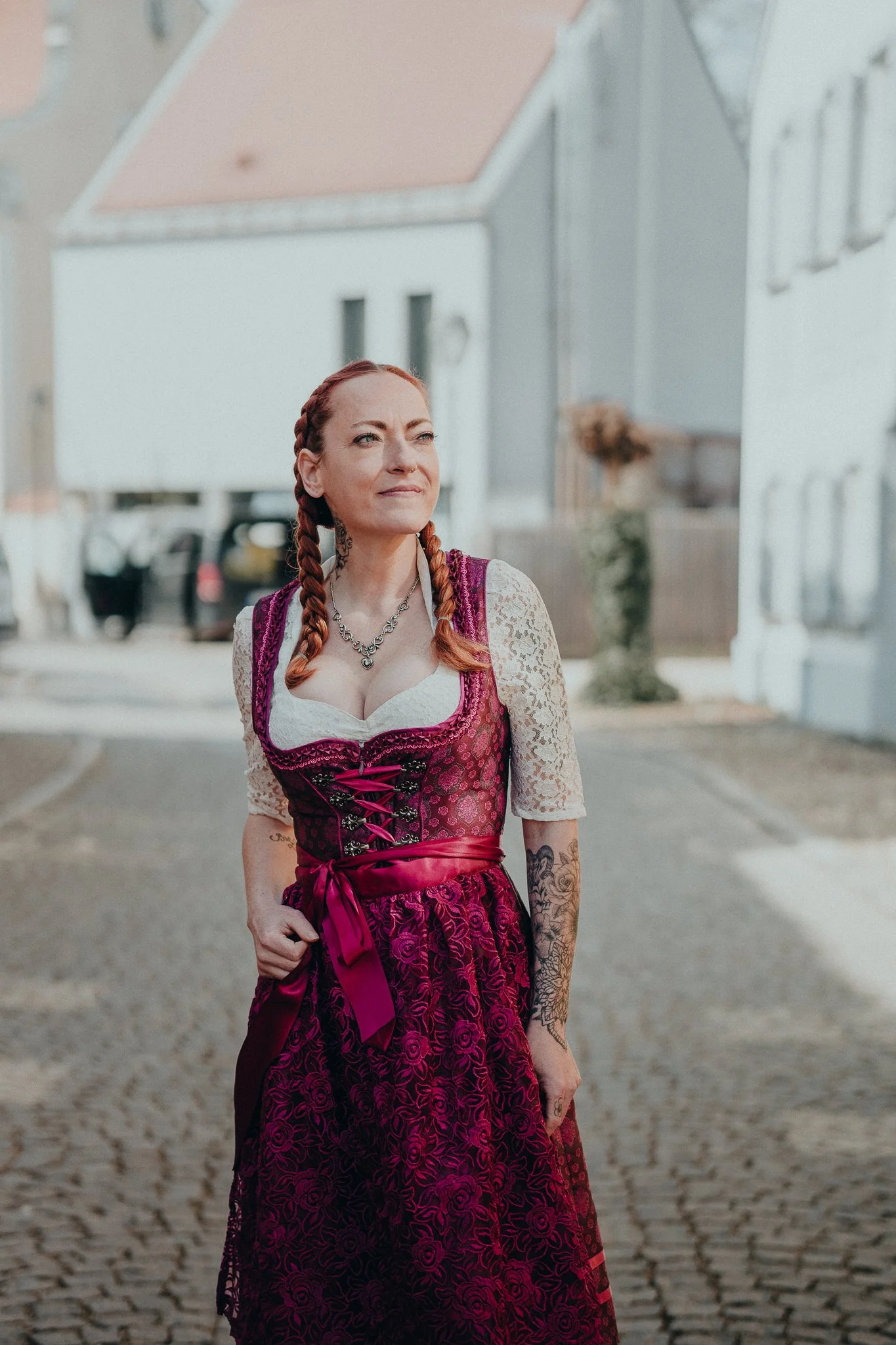 Eine Frau in einem traditionellen Weihnachtsdirndl, mit roten Haaren, auf einer gepflasterten Straße in einer Wohngegend mit weißen Häusern.