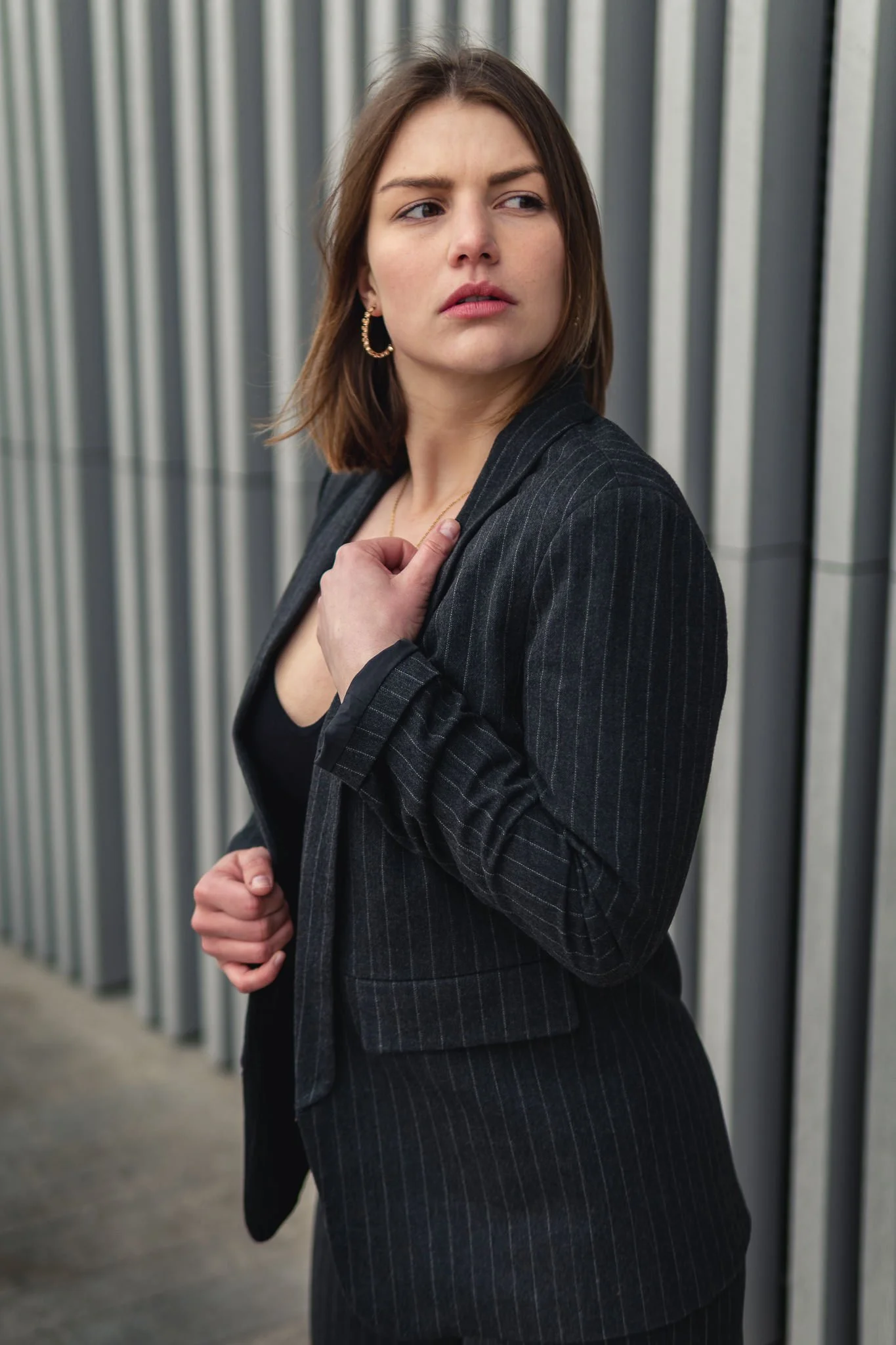 Eine junge Frau mit Schulterlänge braunen Haaren, dunkler Kleidung und Ohrringen steht vor einer vertikal gestreiften Metallwand. Sie blickt nach links mit nachdenklichem Ausdruck.