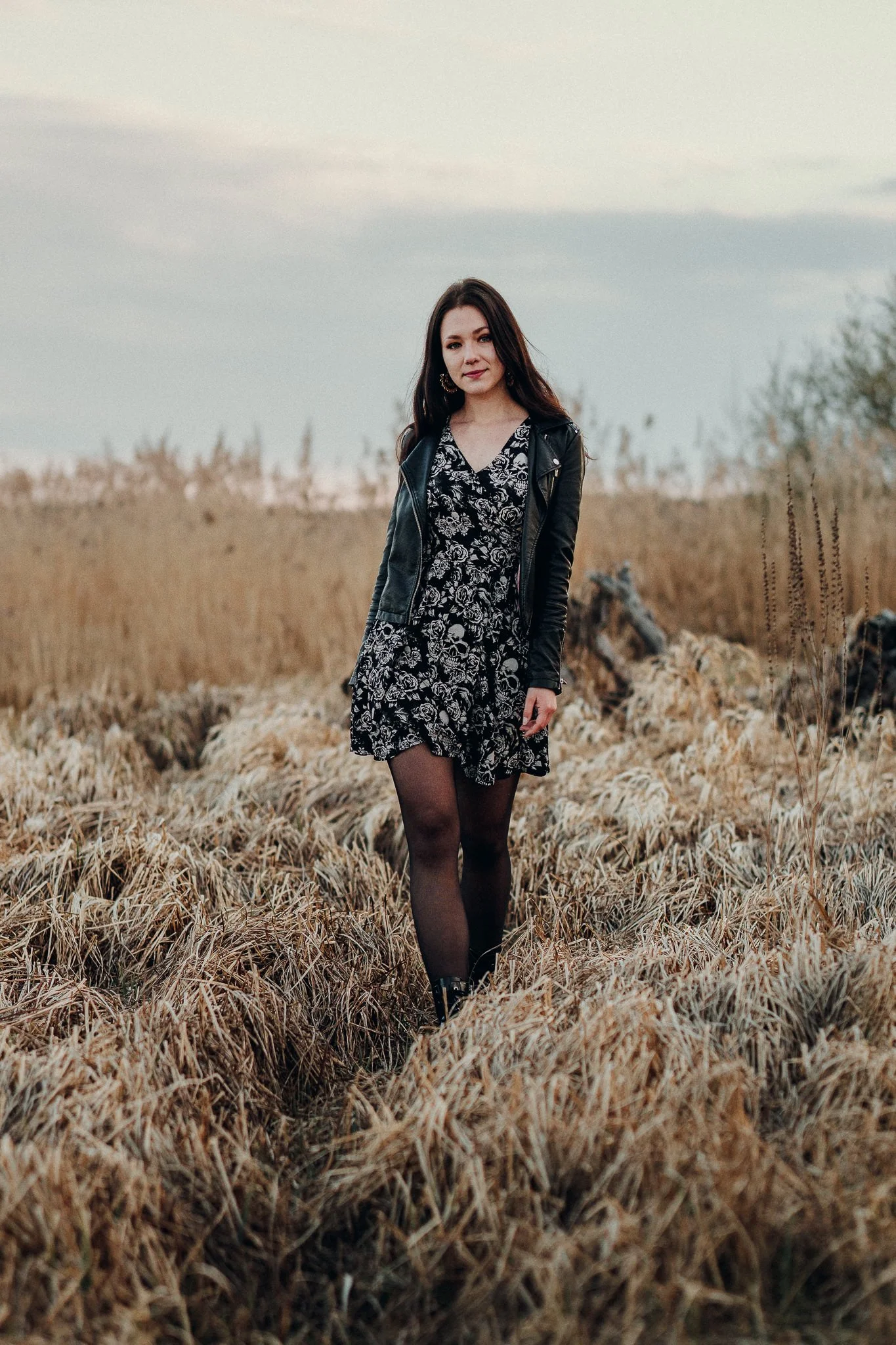 Eine junge Frau steht in einem trockenen Grasfeld bei Sonnenuntergang, trägt ein schwarzes Kleid mit weißen Blumen, eine schwarze Lederjacke und schwarze Strumpfhosen.