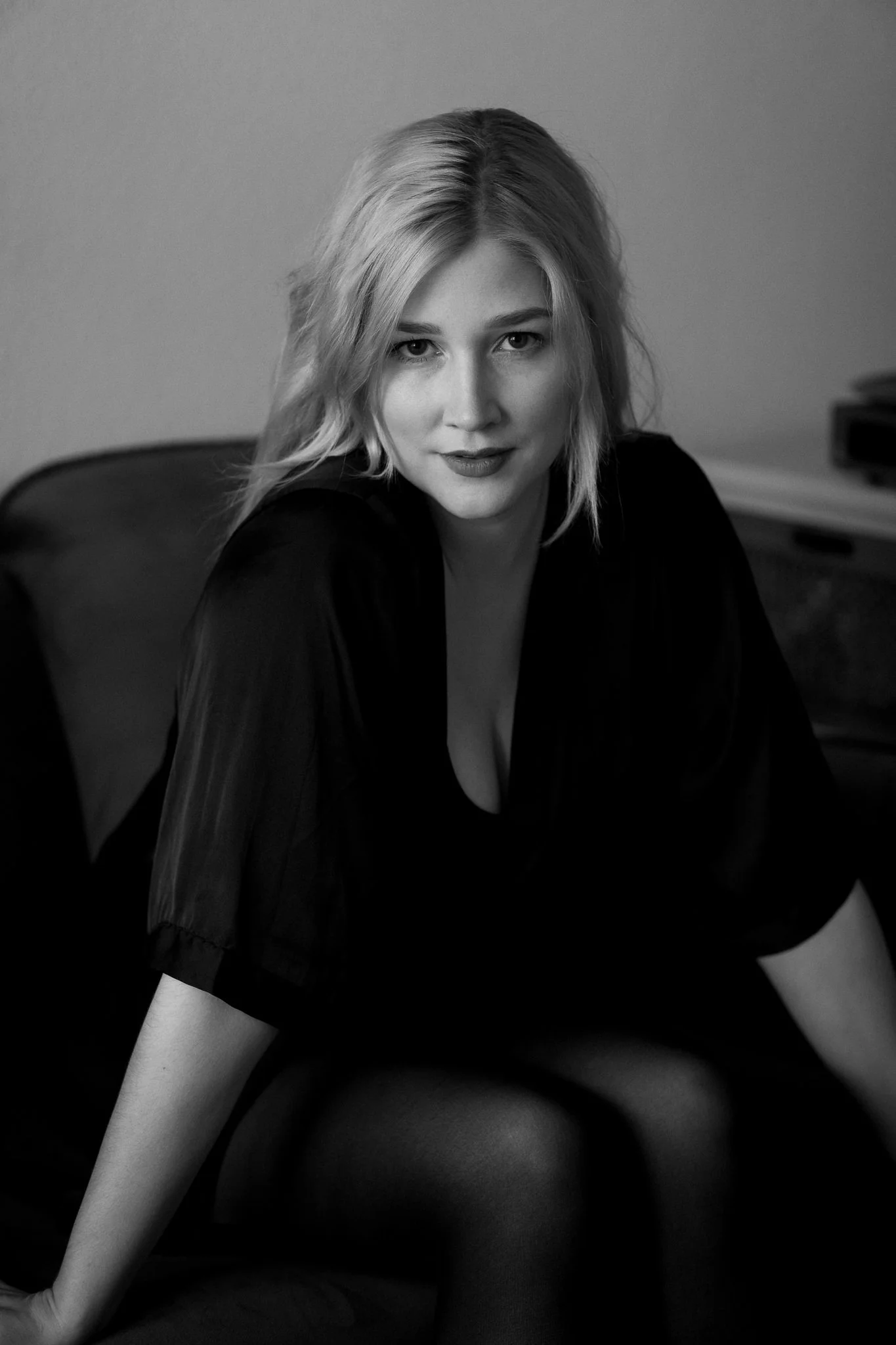 Schwarz-weiß-Porträt einer jungen Frau mit Schulterlangem Haar, sitzend auf einem Stuhl, blickt direkt in die Kamera, trägt dunkle Kleidung, im Hintergrund ein Raum mit regalen.