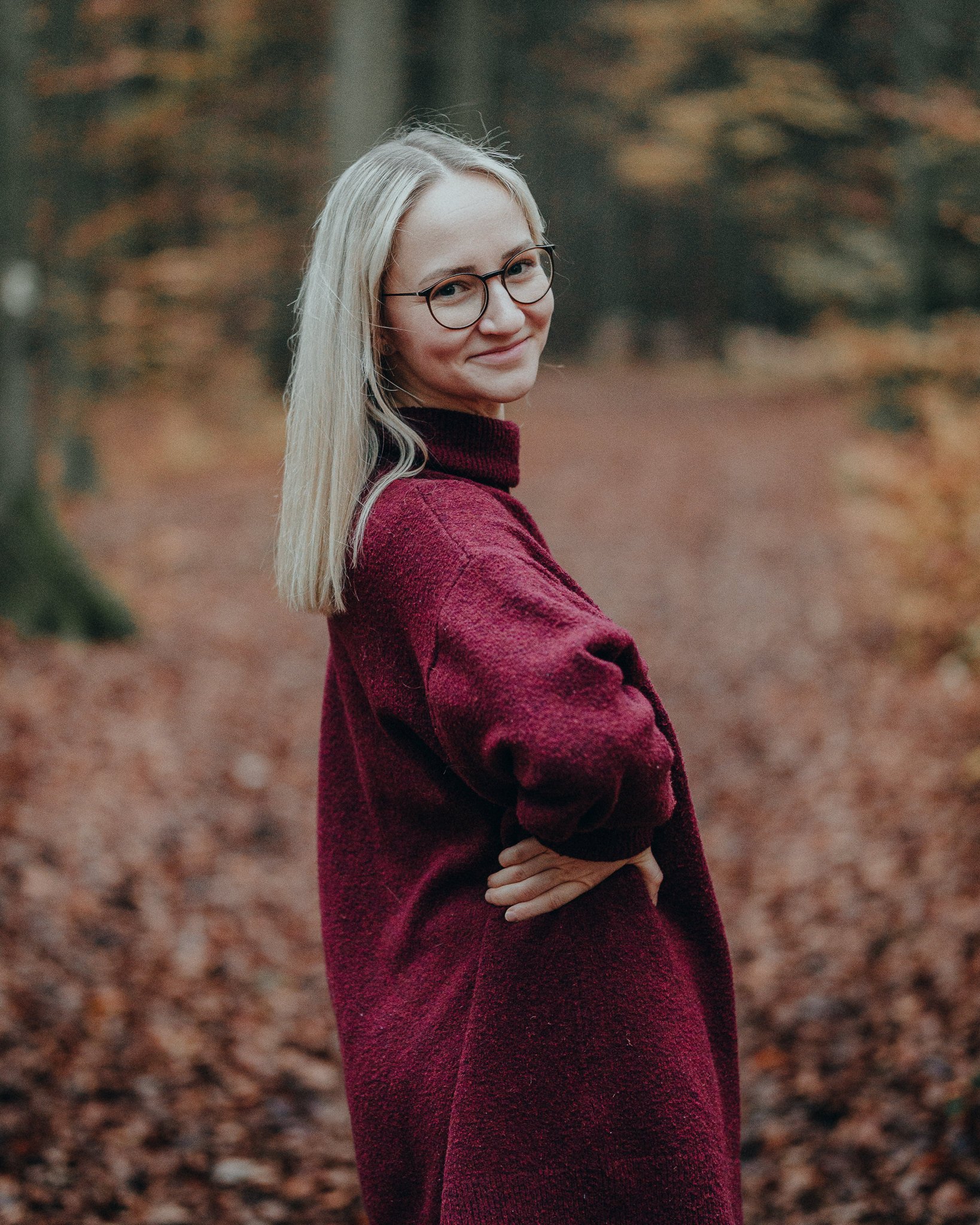 Eine lachende junge Frau mit Brille trägt einen roten Pullover und steht in einem herbstlichen Wald.