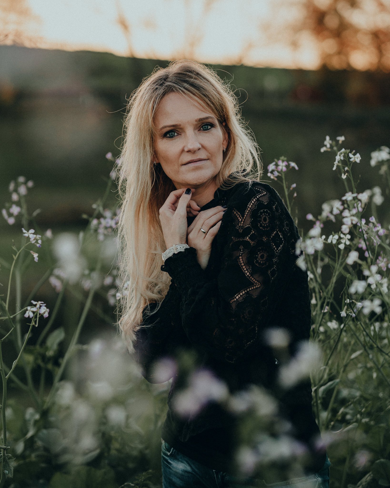 Eine blonde Frau in einem schwarzen Spitzenshirt sitzt in einer Blumenwiese bei Sonnenuntergang.