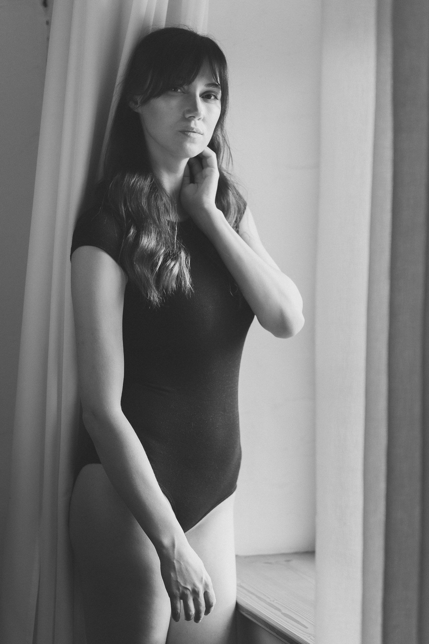 Schwarz-Weiß-Foto einer Frau in einem schwarzen Body, die an einer Wand steht, mit langem, gewelltem Haar und nachdenklichem Blick.