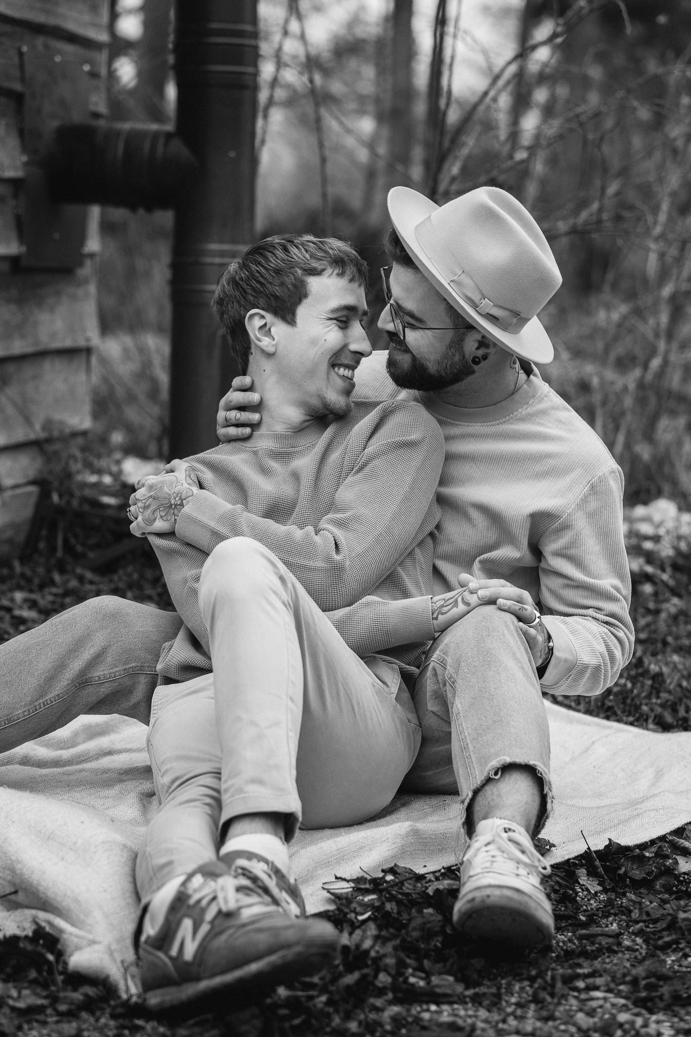 Ein Mann und eine Frau sitzen draußen auf einer Decke, lachen und schauen sich liebevoll an, während sie sich umarmen, in einem Wald oder Garten, schwarz-weiß