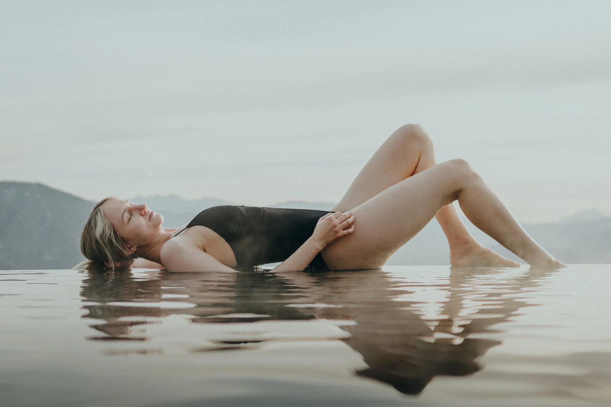 Frau liegt auf dem Rücken im Wasser in einer schwarzen Badebekleidung, mit geschlossenen Augen, umgeben von einer ruhigen Gewässer-Umgebung, Berge im Hintergrund, während sie entspannt in der Natur liegt.