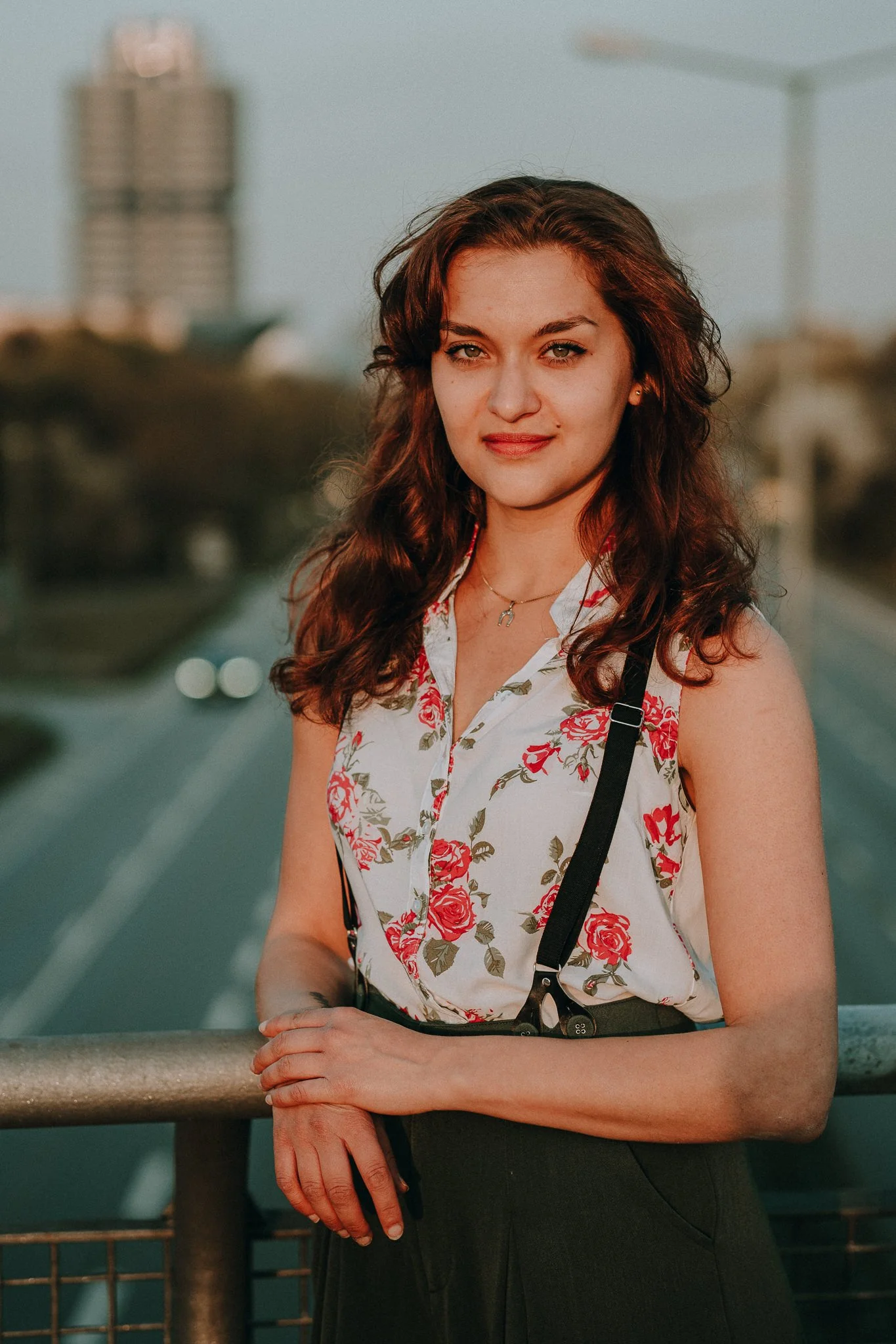 Junge Frau mit braunen, lockigen Haaren, blassem Haut, trägt ein ärmelloses weißes Hemd mit rosenmustern und schwarze Hose, steht auf einer Brücke im Abendlicht, im Hintergrund Stadt und Straße sichtbar.