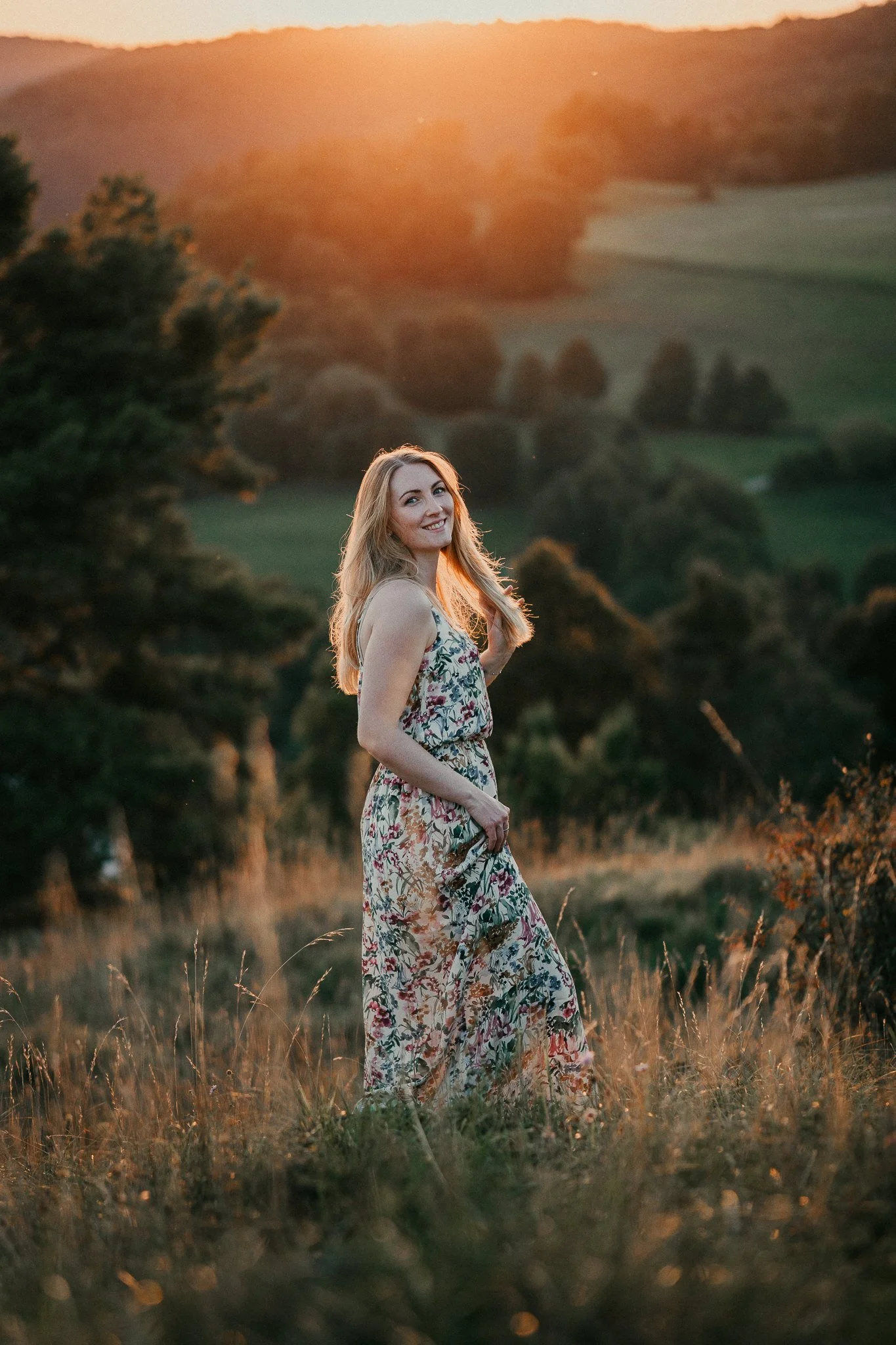 Eine lächelnde junge Frau in einem langestrickten, floral gemusterten Kleid steht im Sonnenuntergang auf einer Wiese mit Hügeln im Hintergrund.