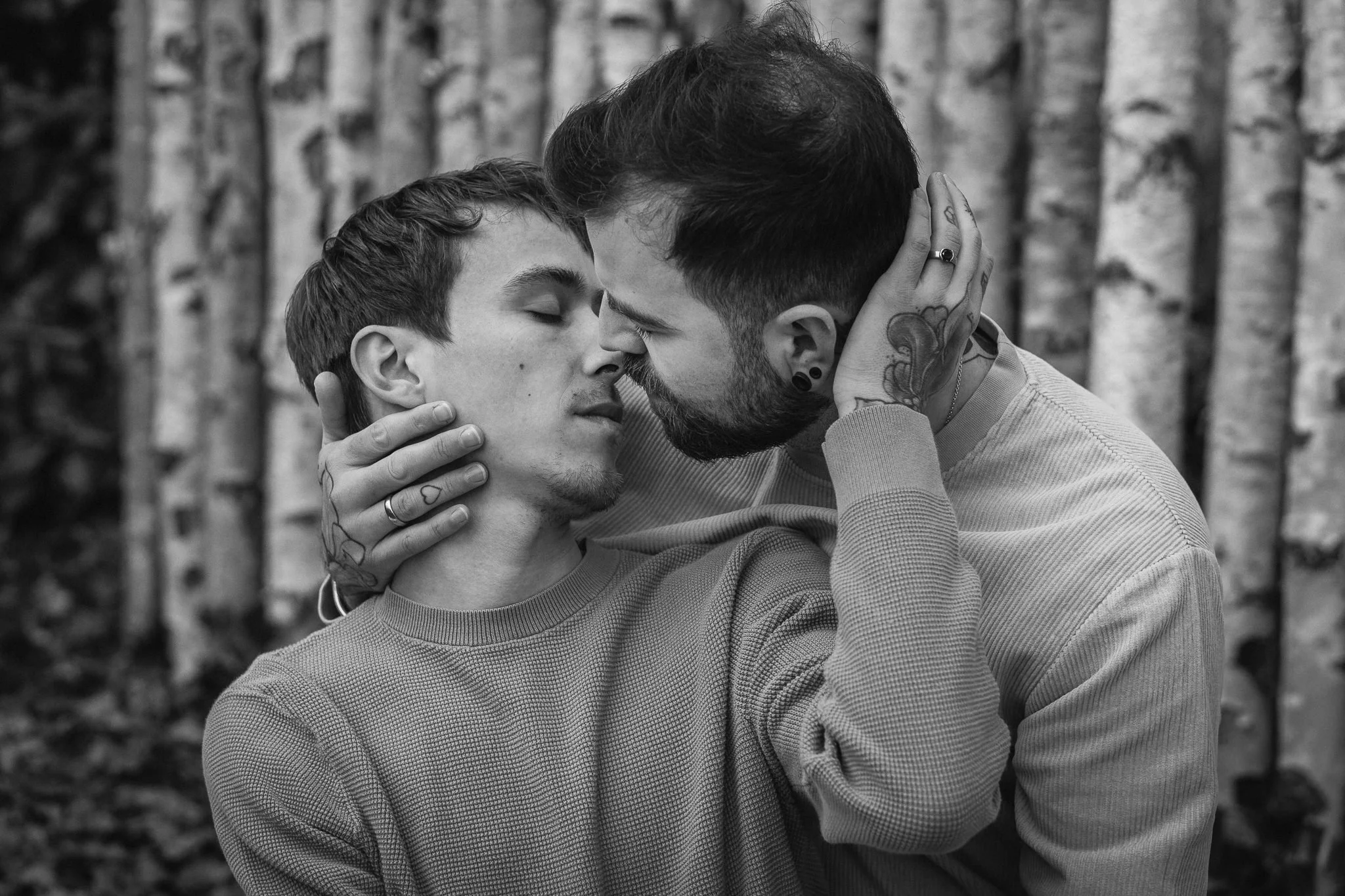 Zwei Männer küssen sich liebevoll, einer hält den Kopf des anderen, im Hintergrund Bäume.