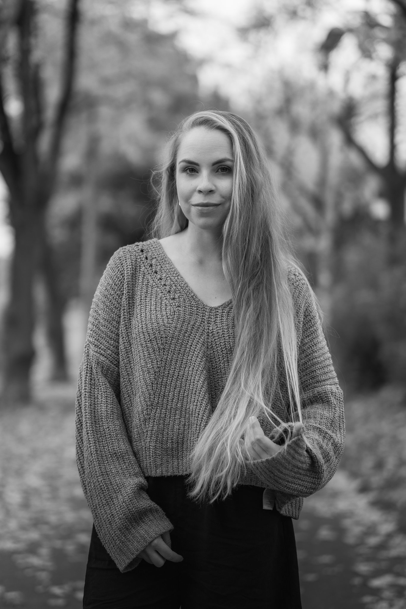 Ein Schwarz-Weiß-Foto einer jungen Frau mit langen, welligen Haaren, die vor Bäumen in einem Park steht, lächelnd in die Kamera blickt, trägt einen Strickpullover.