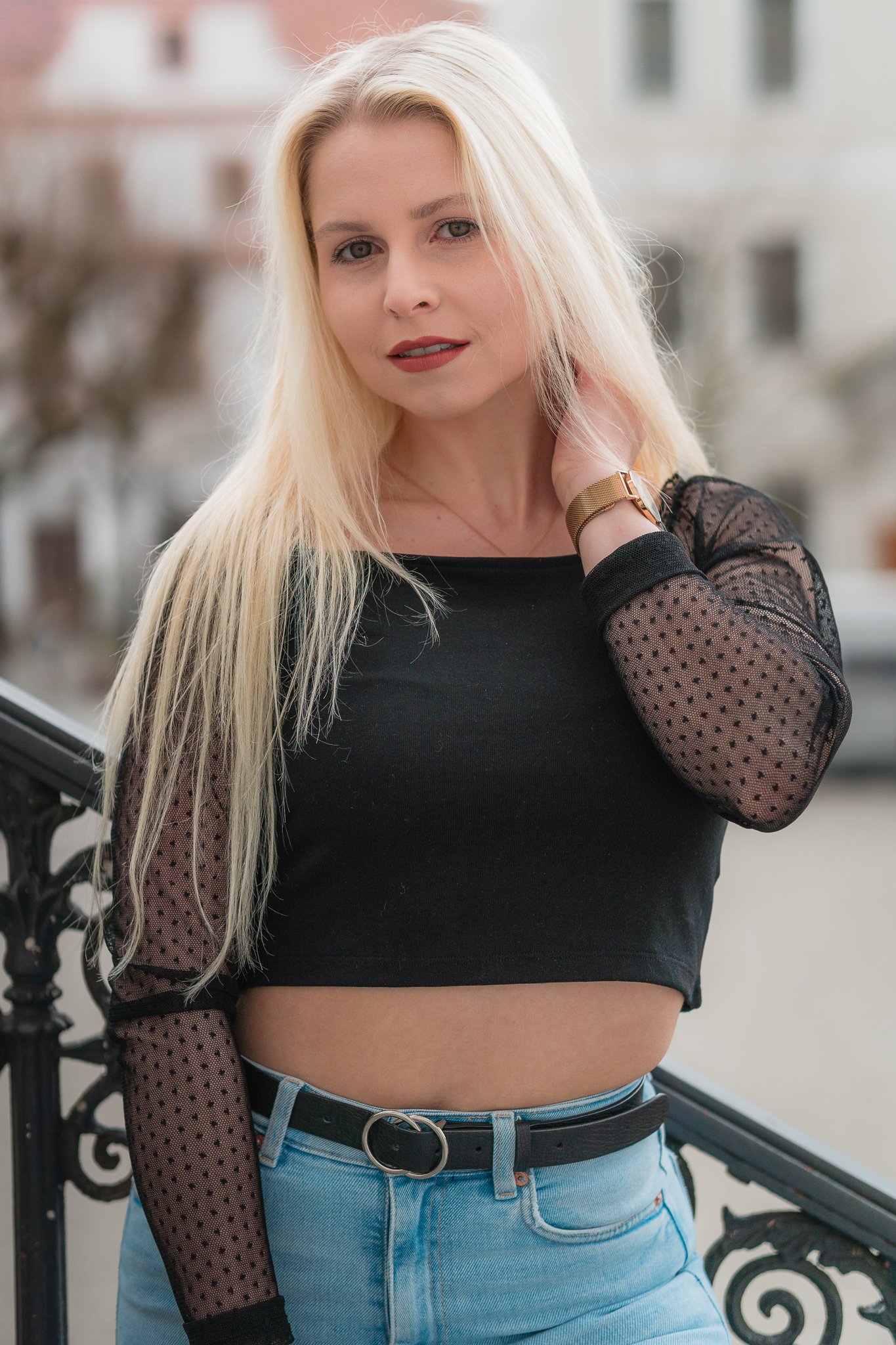 Eine junge blonde Frau steht draußen an einem Geländer und trägt ein schwarzes Top mit durchsichtigen, gepunkteten langen Ärmel und hellblaue Jeans.