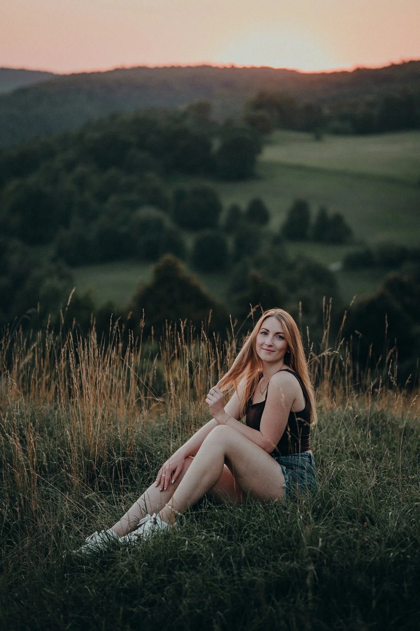 Eine junge Frau sitzt auf einer Wiese bei Sonnenuntergang, umgeben von hohen Gräsern, vor einer hügeligen Landschaft mit Bäumen und einem untergehenden Himmel.