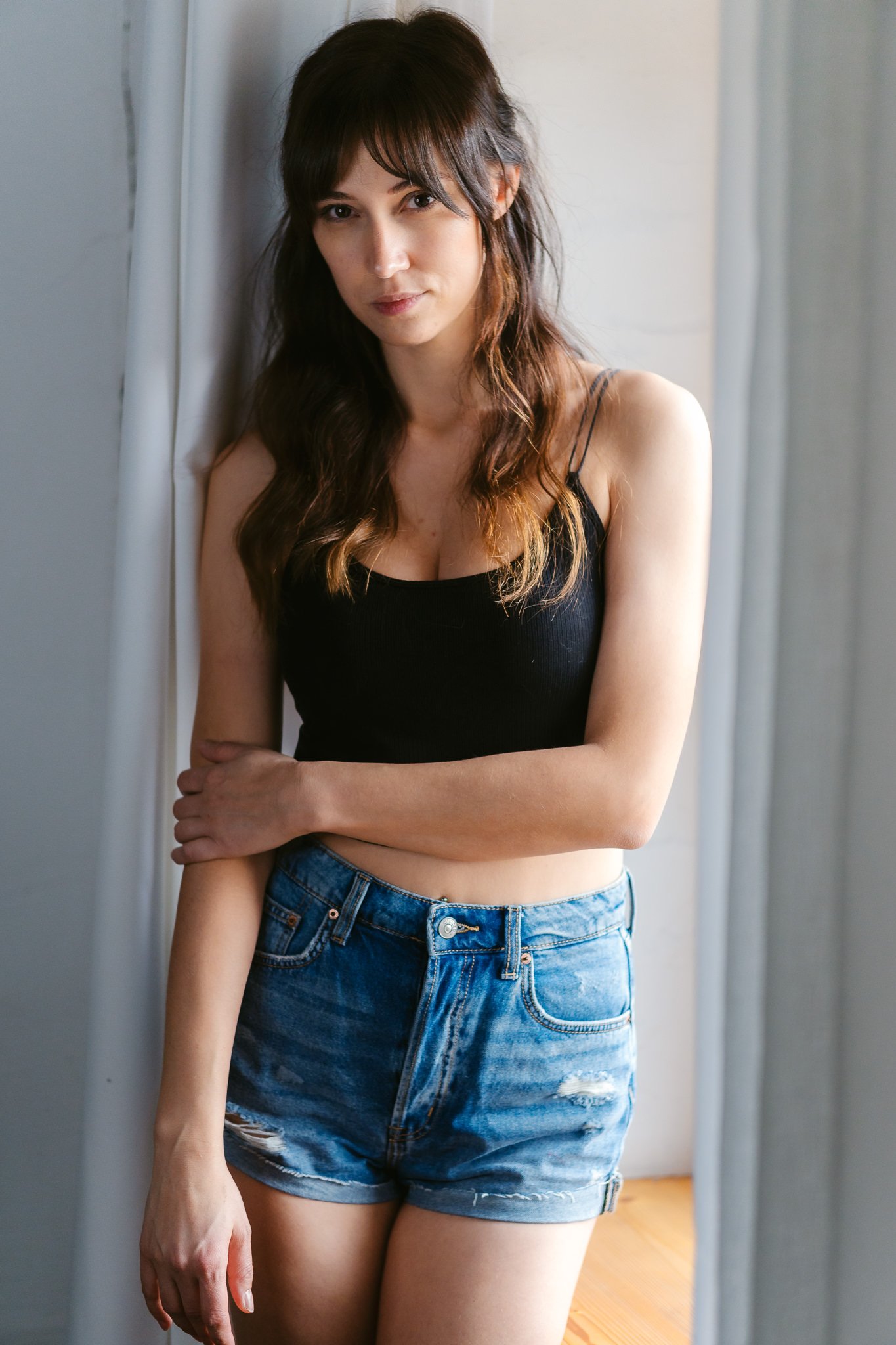 Eine junge Frau mit braunen, gewellten Haaren trägt eine schwarze Top und blue denim Shorts, steht neben einem Vorhang in einem Raum mit natürlichem Licht.