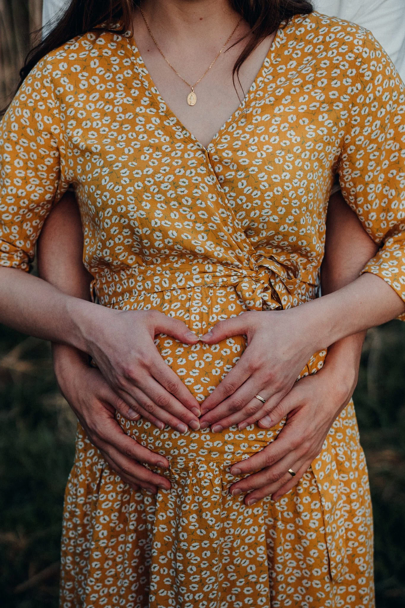 Zwei Frauen umarmen sich und formen ein Herz mit ihren Händen auf einem hervorgehobenen Bauch. Beide tragen Ringe an den Fingern, und die Frau in der Mitte trägt ein gelbes Kleid mit weißen Punkten und einem Rei�verschluss.