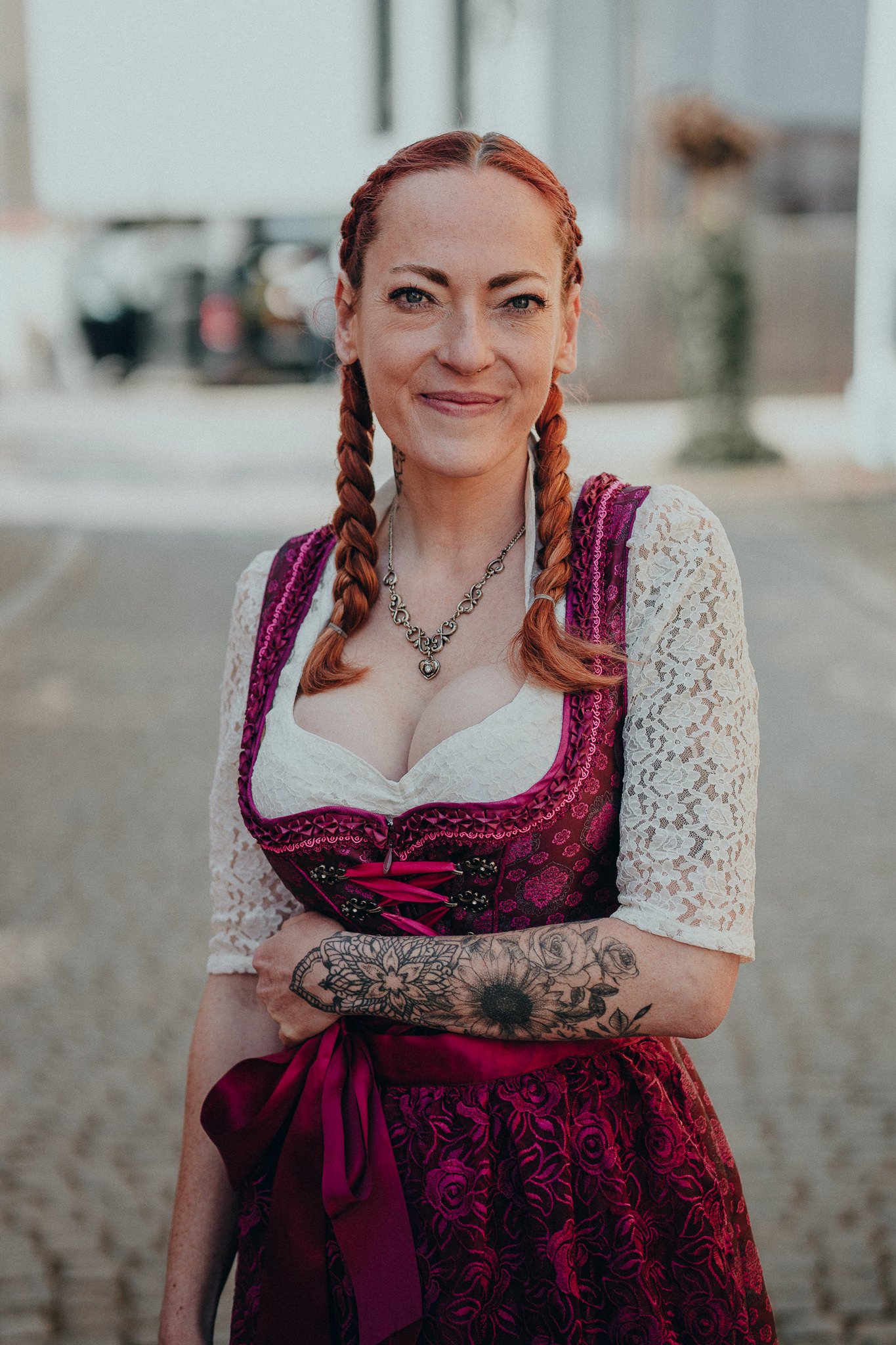 Eine Frau mit roten Zöpfen trägt ein traditionelles Dirndl mit viktorianischem Muster, einer weißen Spitzenbluse und einer Halskette. Sie steht draußen und lächelt.