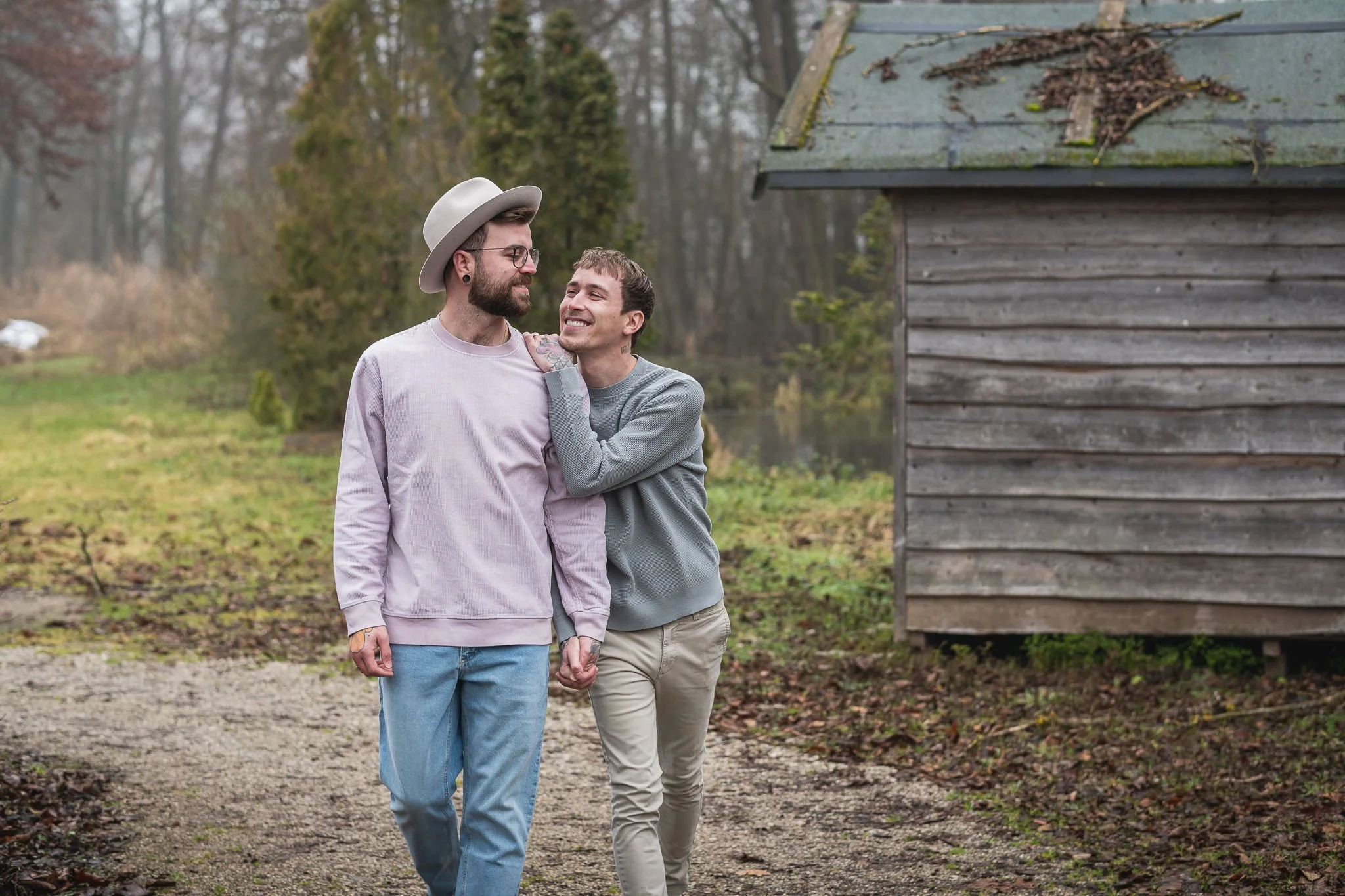 Ein Paar hält Händchen und lacht zusammen beim Spaziergang im Freien auf einem Feldweg, umgeben von Bäumen, mit einem kleinen Holzhaus im Hintergrund.