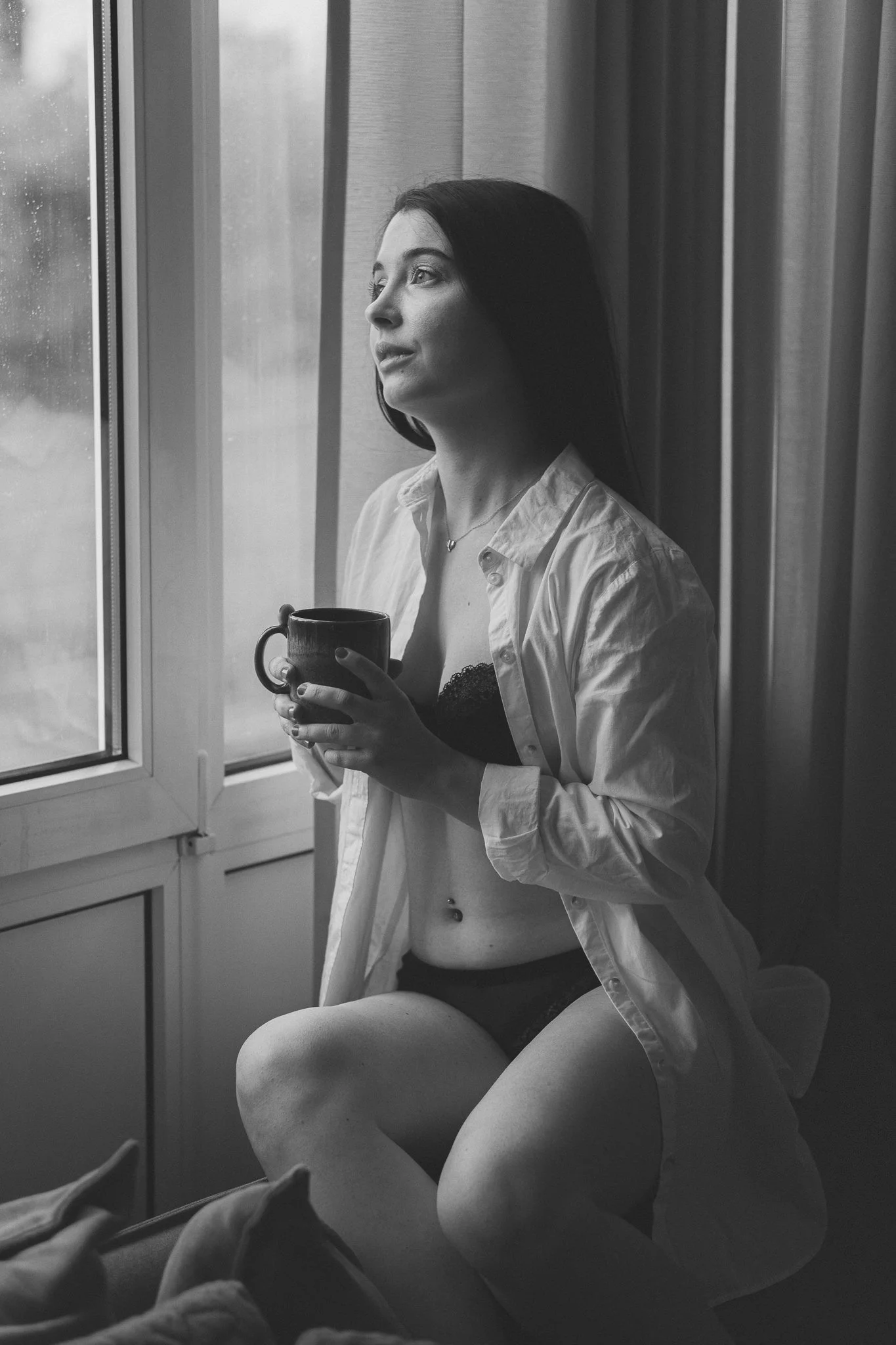 Junge Frau sitzt am Fenster, trägt offen Hemd, Unterwäsche, hält eine Tasse Kaffee, schaut nach draußen, im schwarzen und weißen Bild.