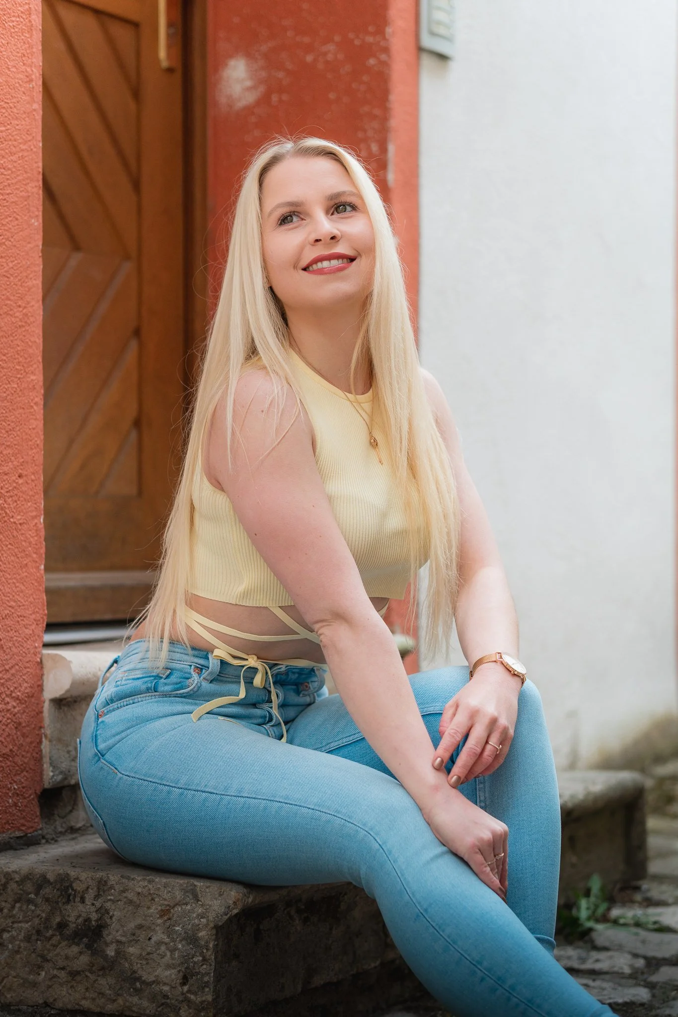 Eine junge blonde Frau sitzt auf einer Steintreppe vor einem roten und weißen Wand. Sie trägt eine gelbe ärmellose Oberteil und blaue Jeans, lächelt leicht und schaut nach oben.