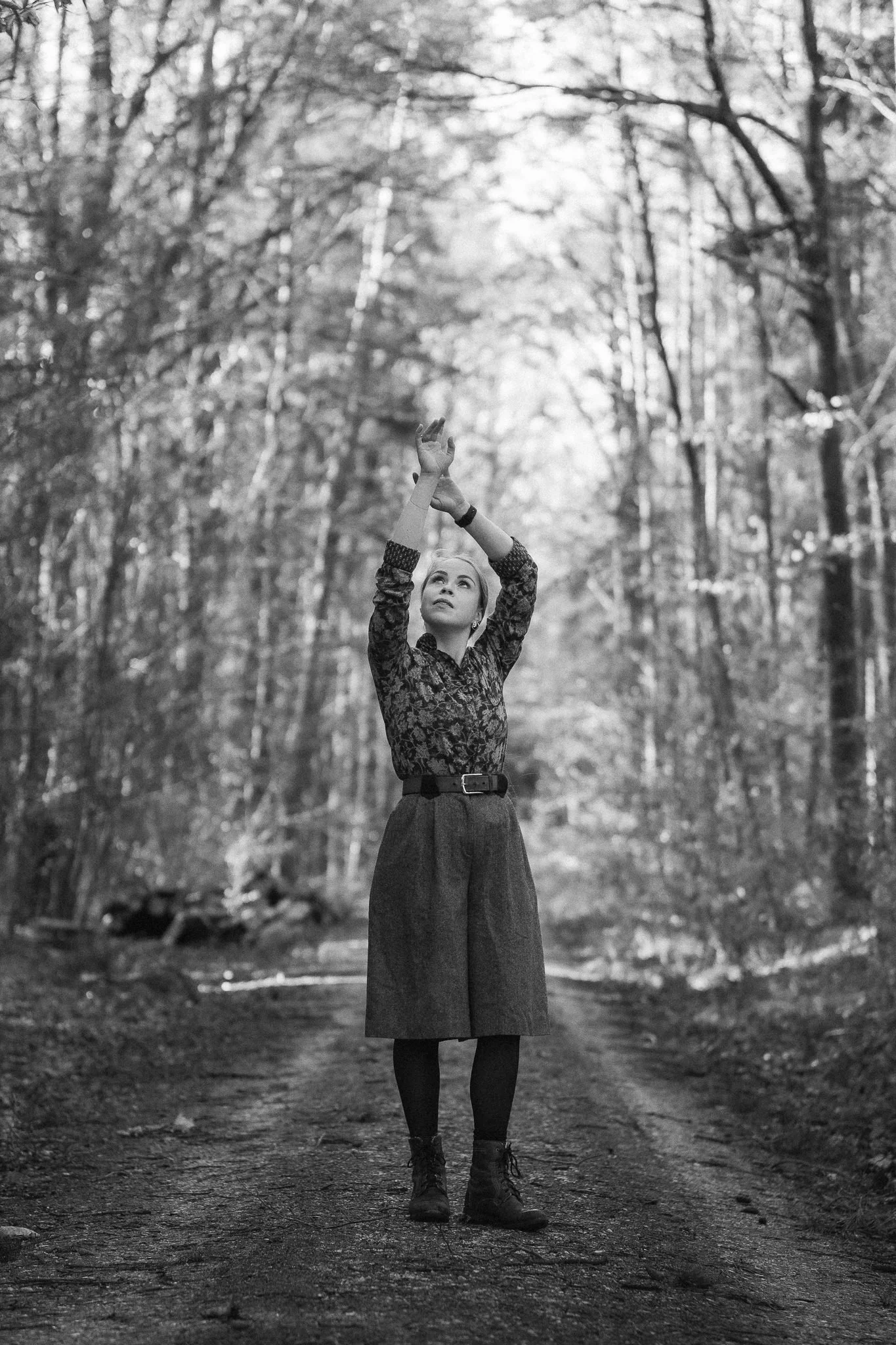 Eine Frau steht auf einem Waldweg und hebt die Arme hoch, um einen Gegenstand zu greifen oder zu berühren. Das Bild ist schwarz-weiß.