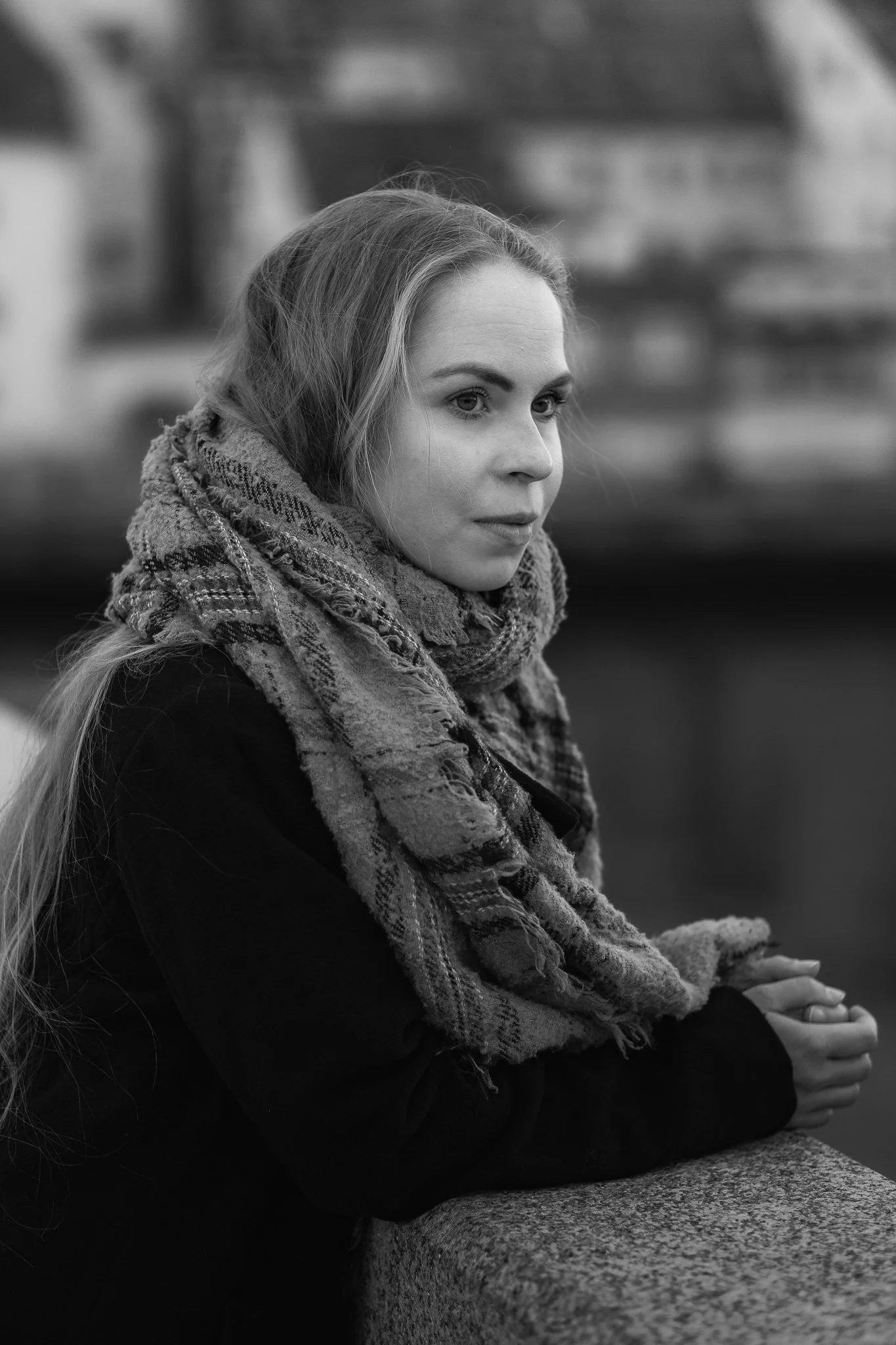 Ein junges Mädchen mit langem Haar, schwarze Jacke und gesteppter Schal sitzt an einer Brüstung, blickt nach rechts, im Hintergrund eine Stadtansicht und Wasser. Schwarz-Weiß-Foto.