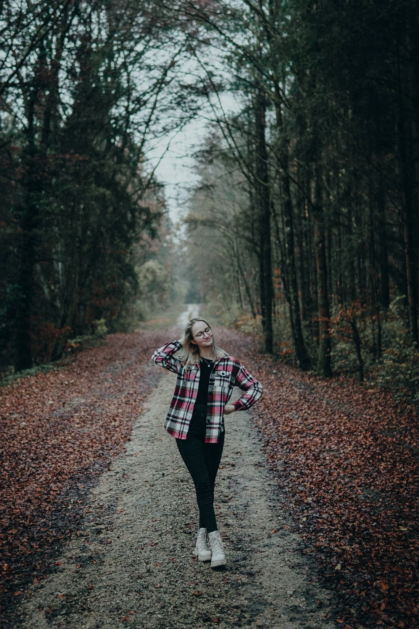 Junge Frau in kariertem Hemd und schwarzen Hosen, die auf einer Waldweg im Herbst steht und die Augen geschlossen hat.