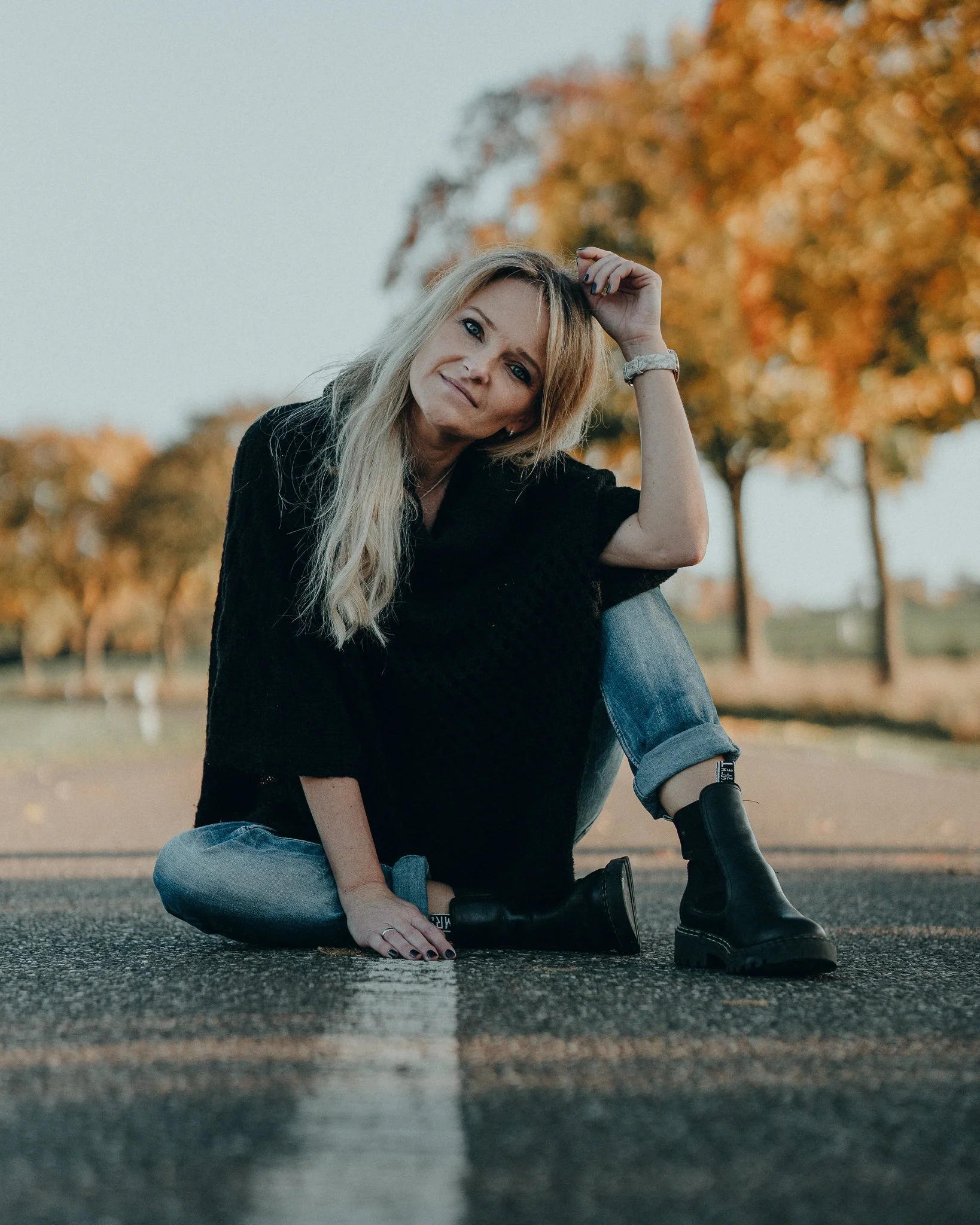 Eine blonde Frau sitzt auf einer Straße im Herbst, trägt dunkle Kleidung und schwarze Stiefel, mit Bäumen im goldenen Laub im Hintergrund.