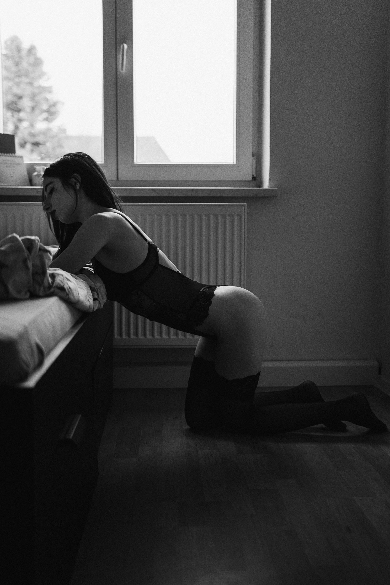 Schwarze und weiße Fotografie einer jungen Frau in Dessous auf Knien auf dem Boden, lehnt sich gegen eine Bettkante, in einem Raum neben einem Fenster.
