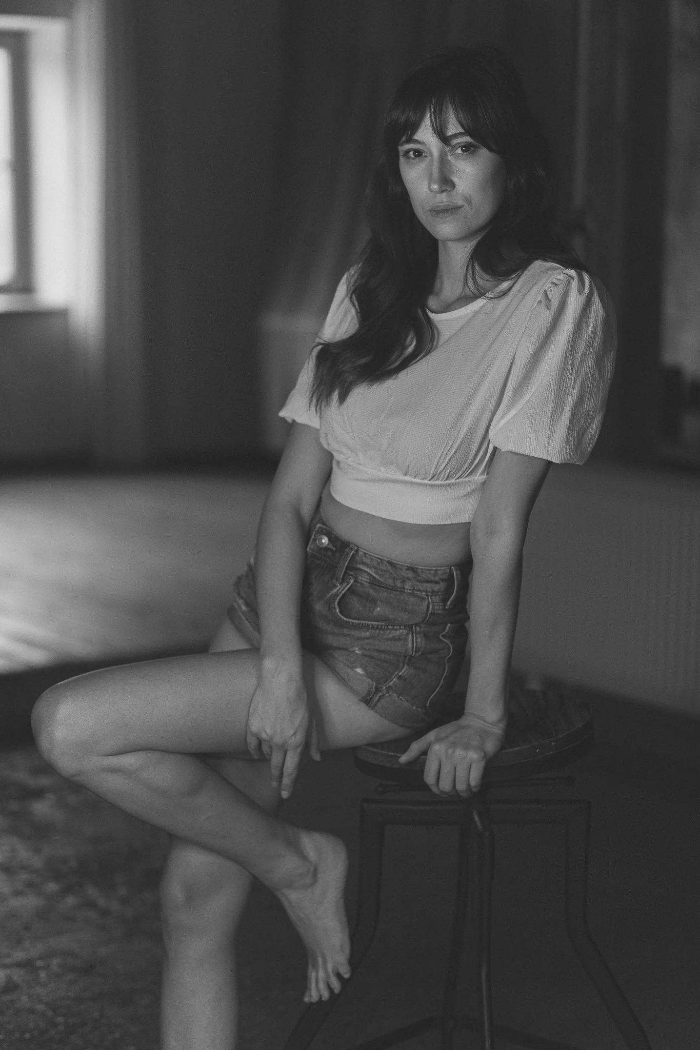Schwarz-Weiß-Foto einer Frau mit dunklen, longen Haaren, die auf einem Stuhl sitzt, ein Bein über das andere geschlagen, in einem Raum mit natürlichem Licht.