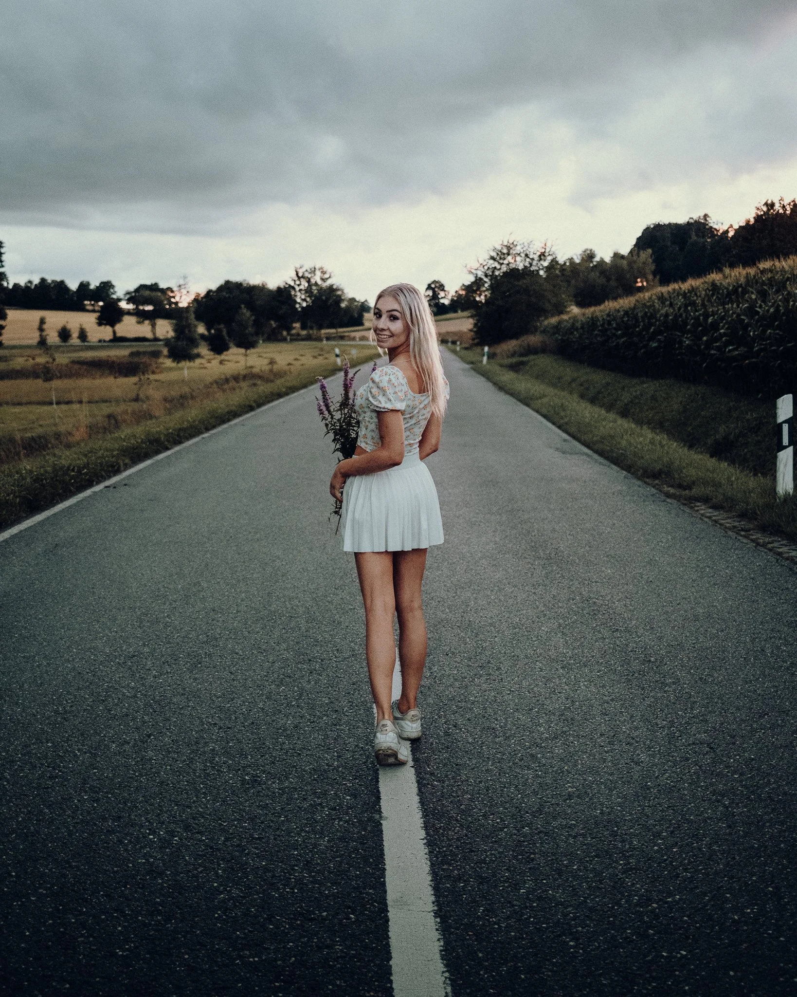 Eine junge Frau mit blonden Haaren trägt ein weißes Kleid und läuft auf einer leeren Landstraße mit dunklem Himmel im Hintergrund, während sie eine Blumenstrauß in der Hand hält und in die Kamera lächelt.