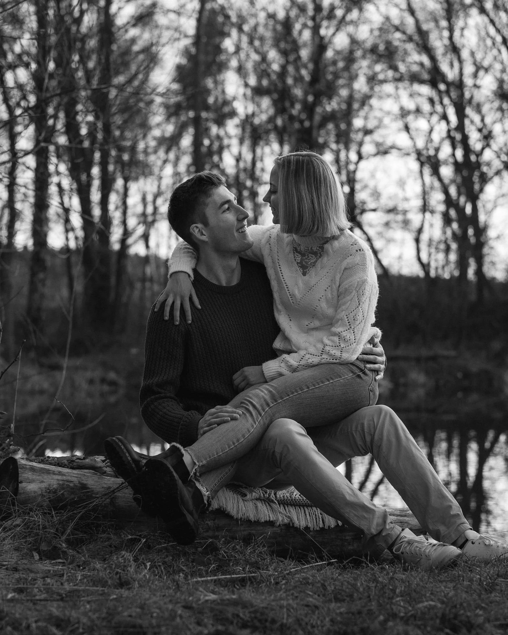 Ein Paar sitzt im Wald, die Frau sitzt auf den Knien des Mannes, beide lachen und schauen sich an, umgeben von Bäumen, bei Sonnenuntergang.