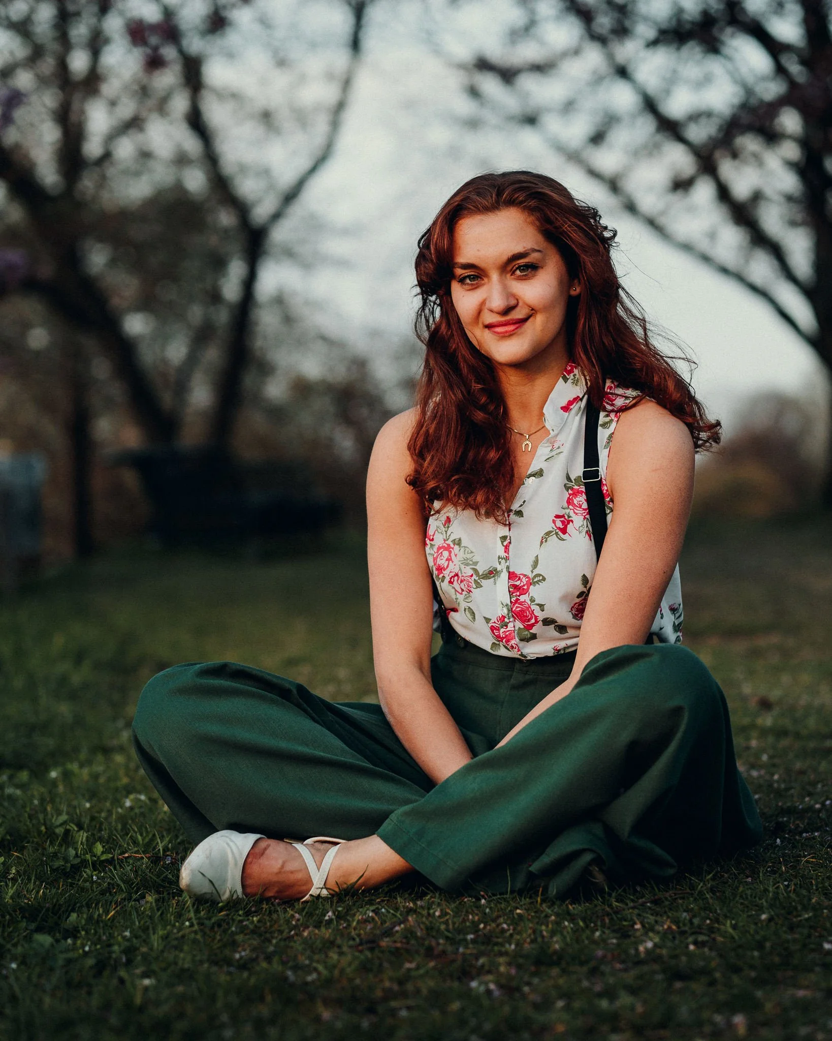 Eine junge Frau sitzt im Freien auf dem Gras, trägt ein weißes ärmelloses Top mit Blumenmuster und grüne Hosen, lächelt und blickt in die Kamera, im Hintergrund sind Bäume und ein bewölkter Himmel.