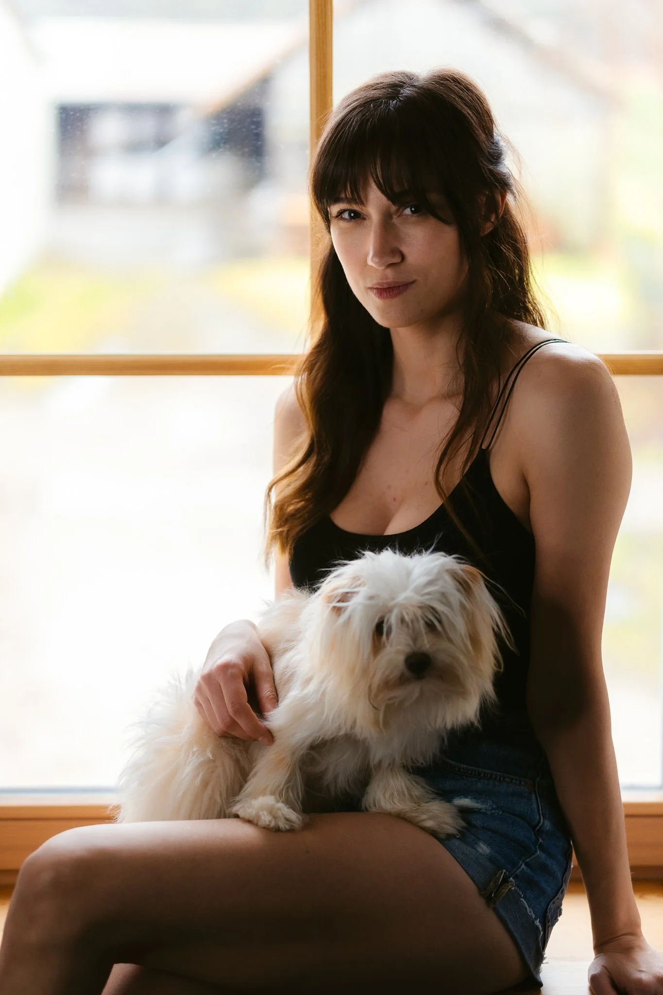Eine junge Frau sitzt vor einem Fenster und hält einen kleinen, weißen, langhaarigen Hund auf dem Schoß.