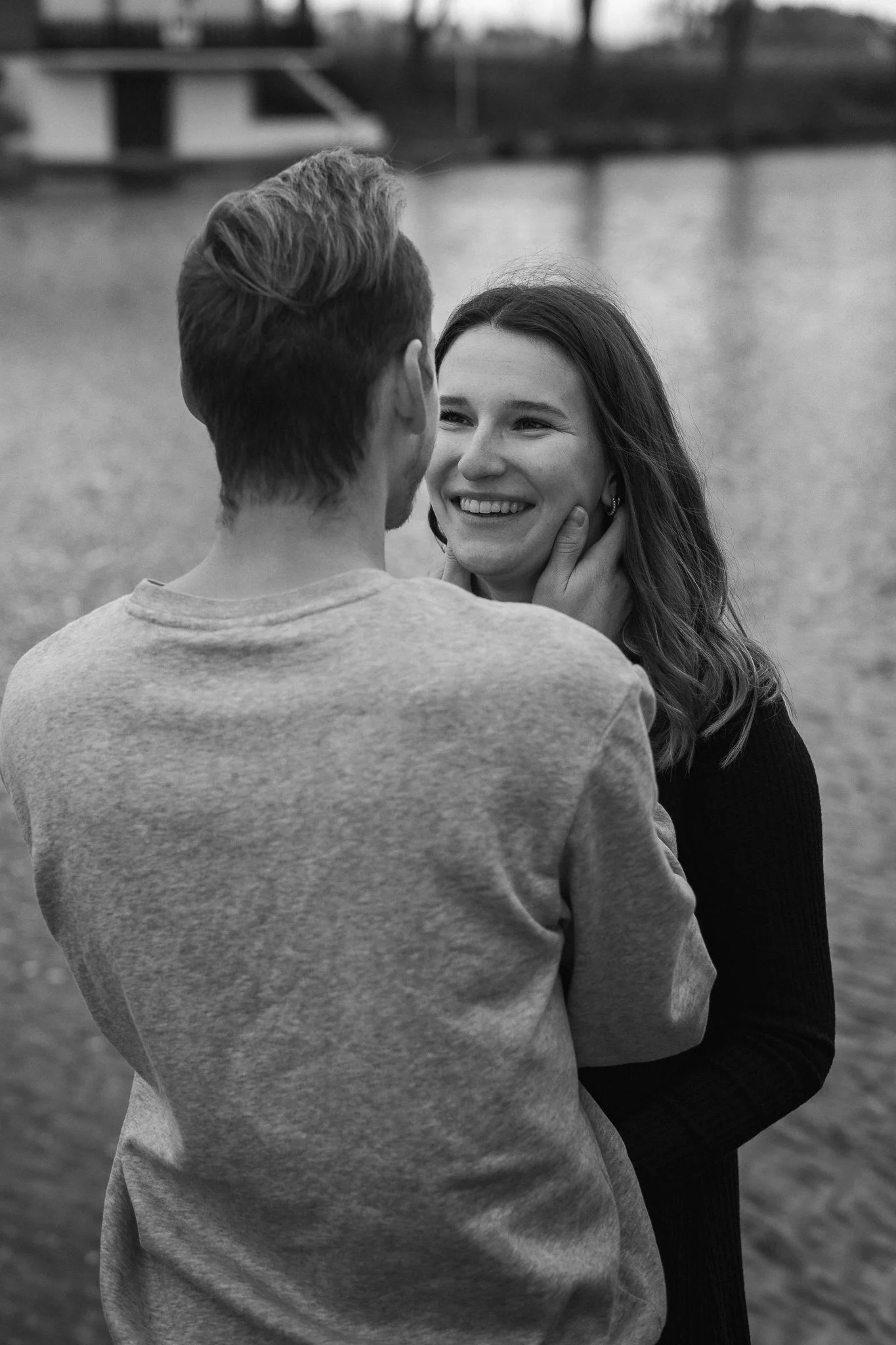 Ein Mann und eine Frau stehen nah beieinander am Wasser, der Mann hält die Frau im Arm, beide lächeln sich an, die Szene ist in Schwarz-Weiß.
