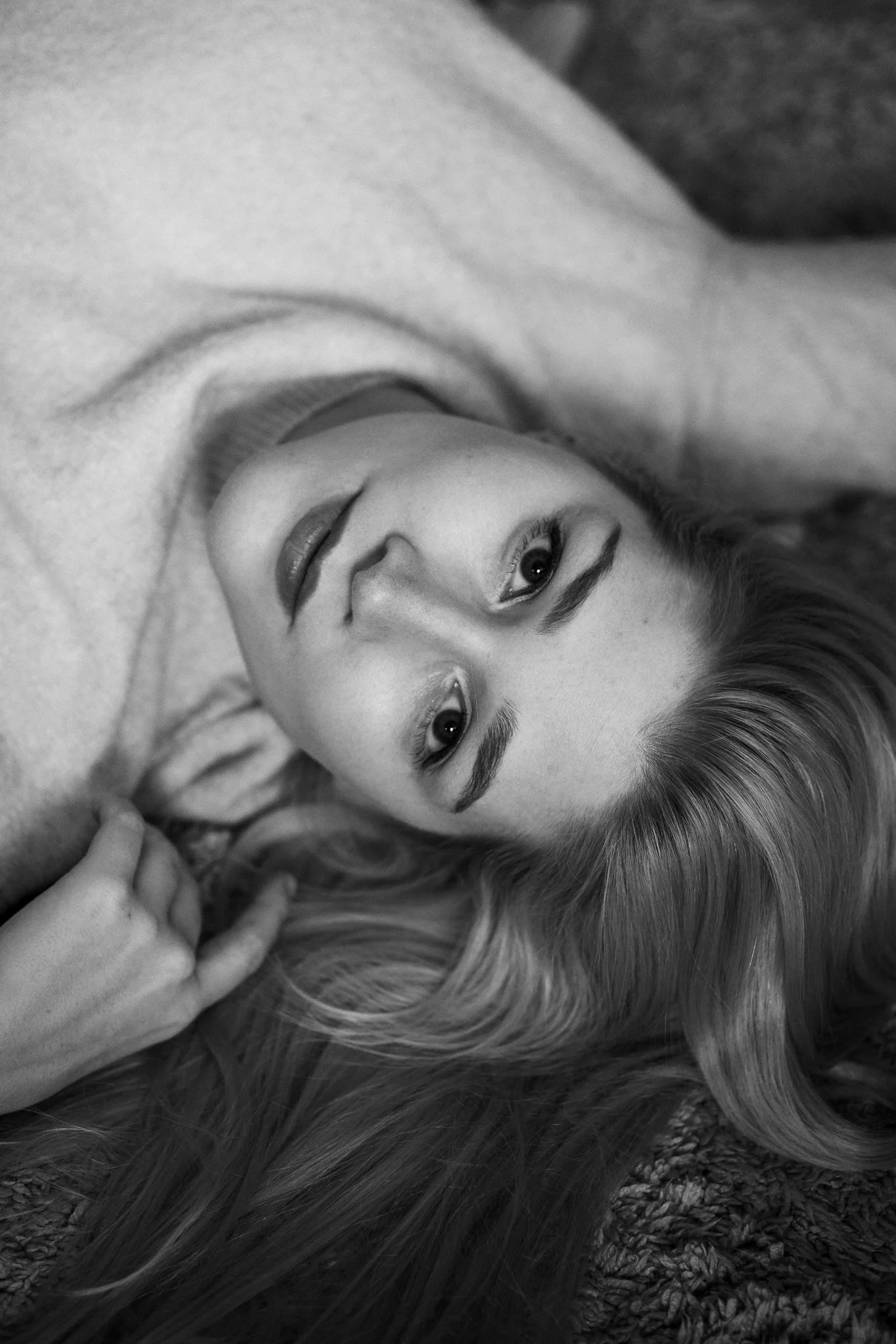 Schwarz-weiß-Fotografie eines jungen Mädchens mit langen Haaren, das auf einer Oberfläche liegt, Blick direkt in die Kamera, entspannt und nachdenklich.