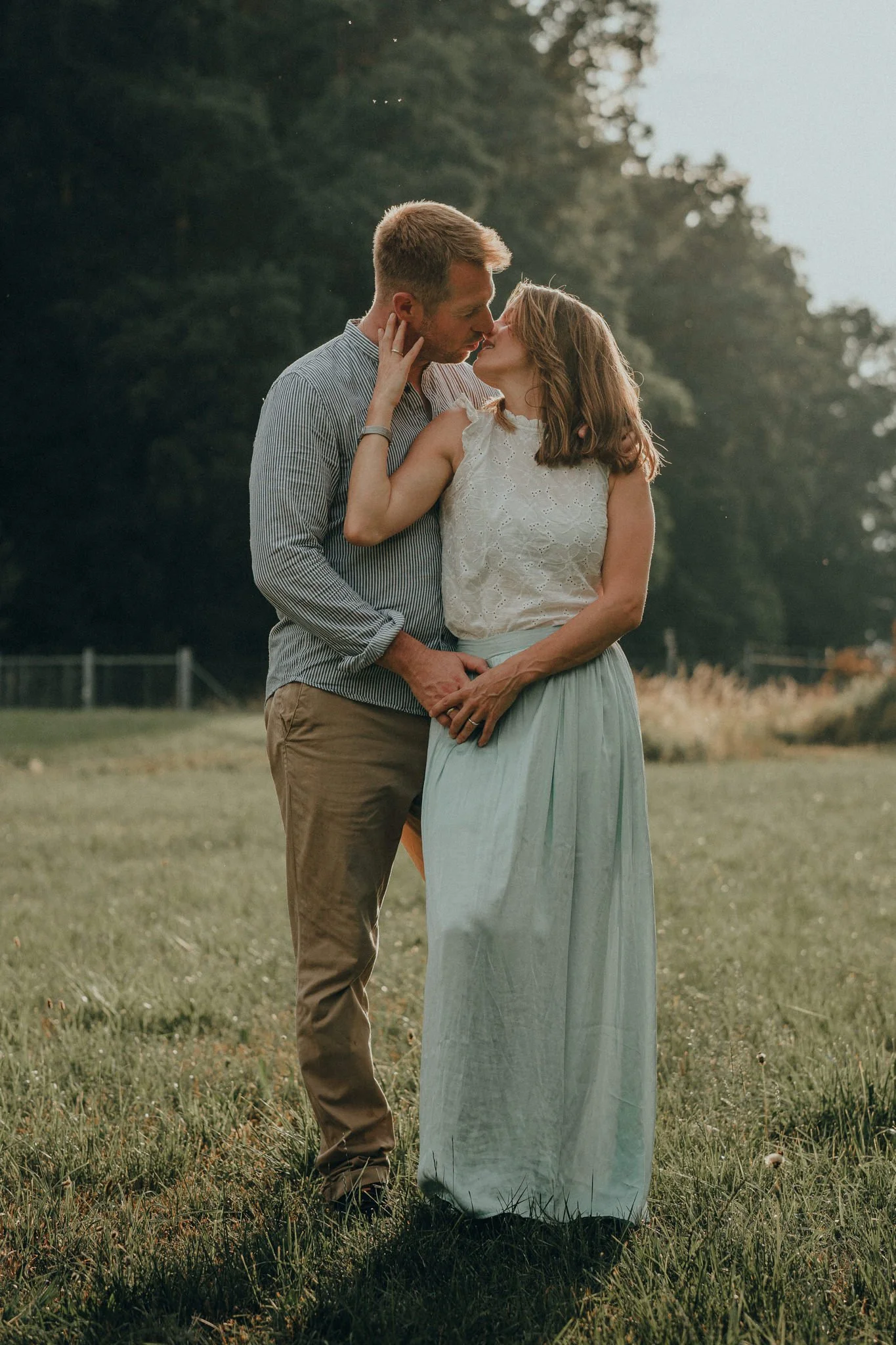 Ein Paar beim Kuscheln im Freien auf einer Wiese bei Sonnenuntergang, umgeben von Bäumen.