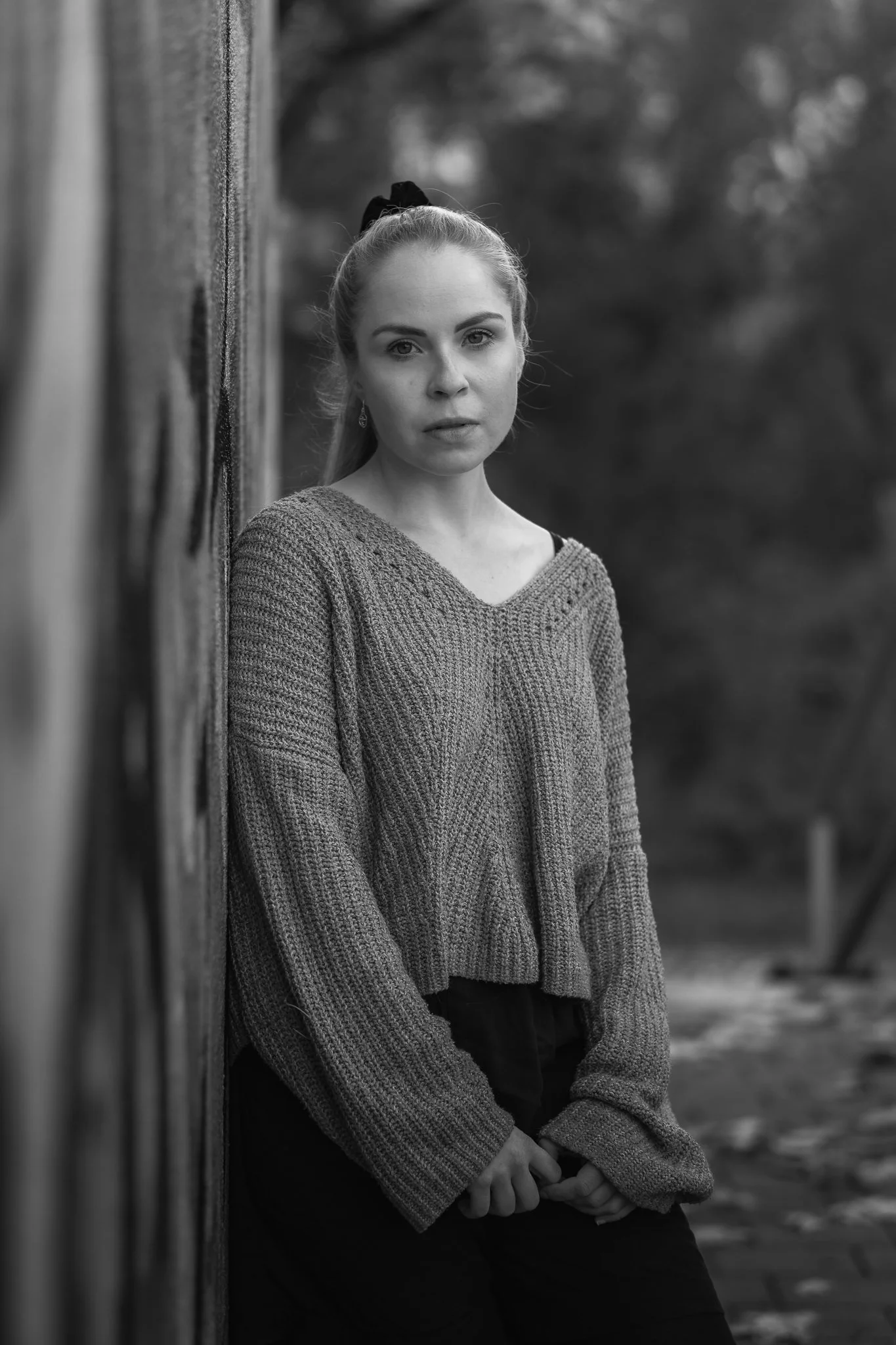 Schwarz-weiß-Fotografie eines jungen Mädchens, das an einer Holzwand lehnt, mit langen Haaren, die zu einem Pferdeschwanz gebunden sind, und in einem gemütlichen Pullover gekleidet ist.