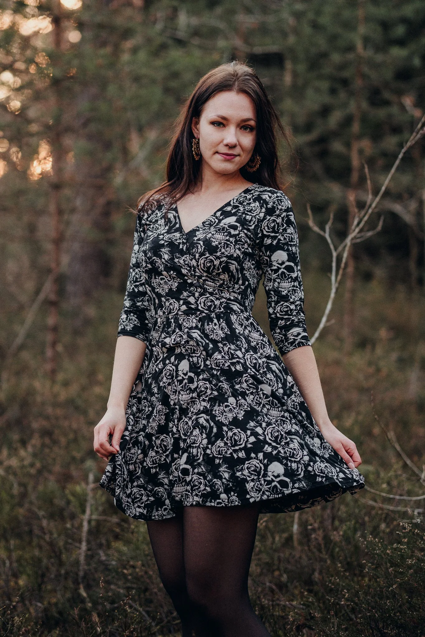 Eine junge Frau steht im Wald, trägt ein schwarzes Kleid mit weißen Totenkopf- und Rosenmustern, schwarze Strumpfhosen, hat braune Haare und trägt goldene Ohrringe.