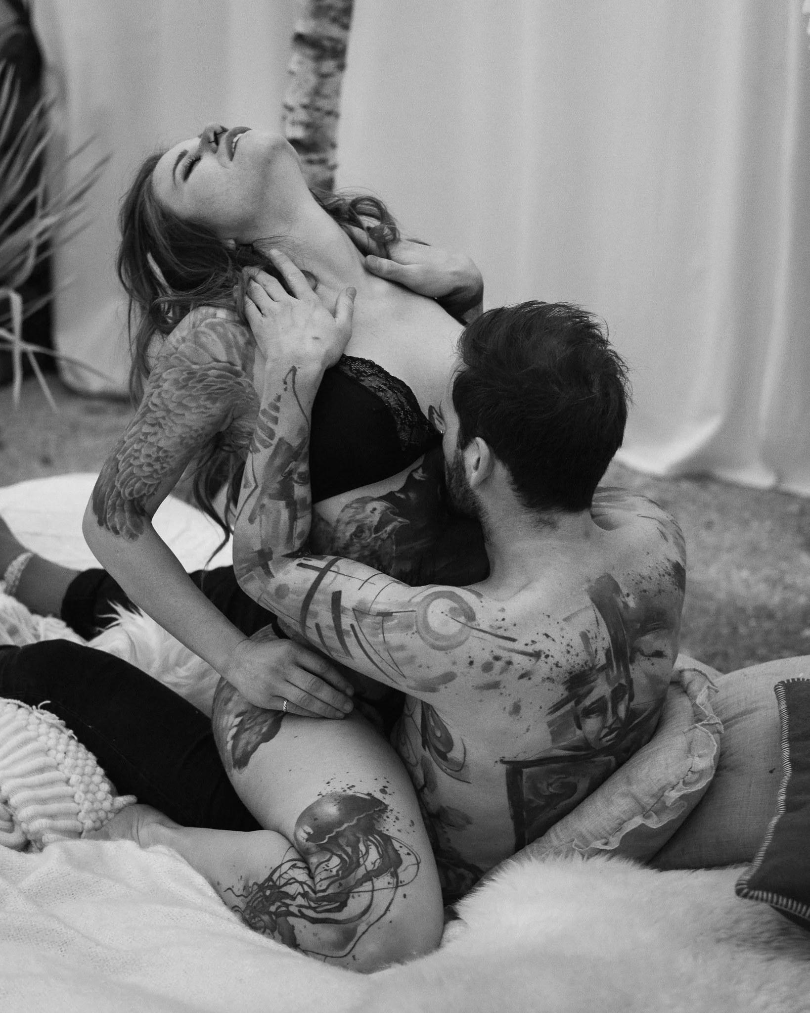 Ein Tattoo-tragendes Paar sitzt auf einem Bett, umarmt sich und zeigt intime Momente, während sie sich ansehen.