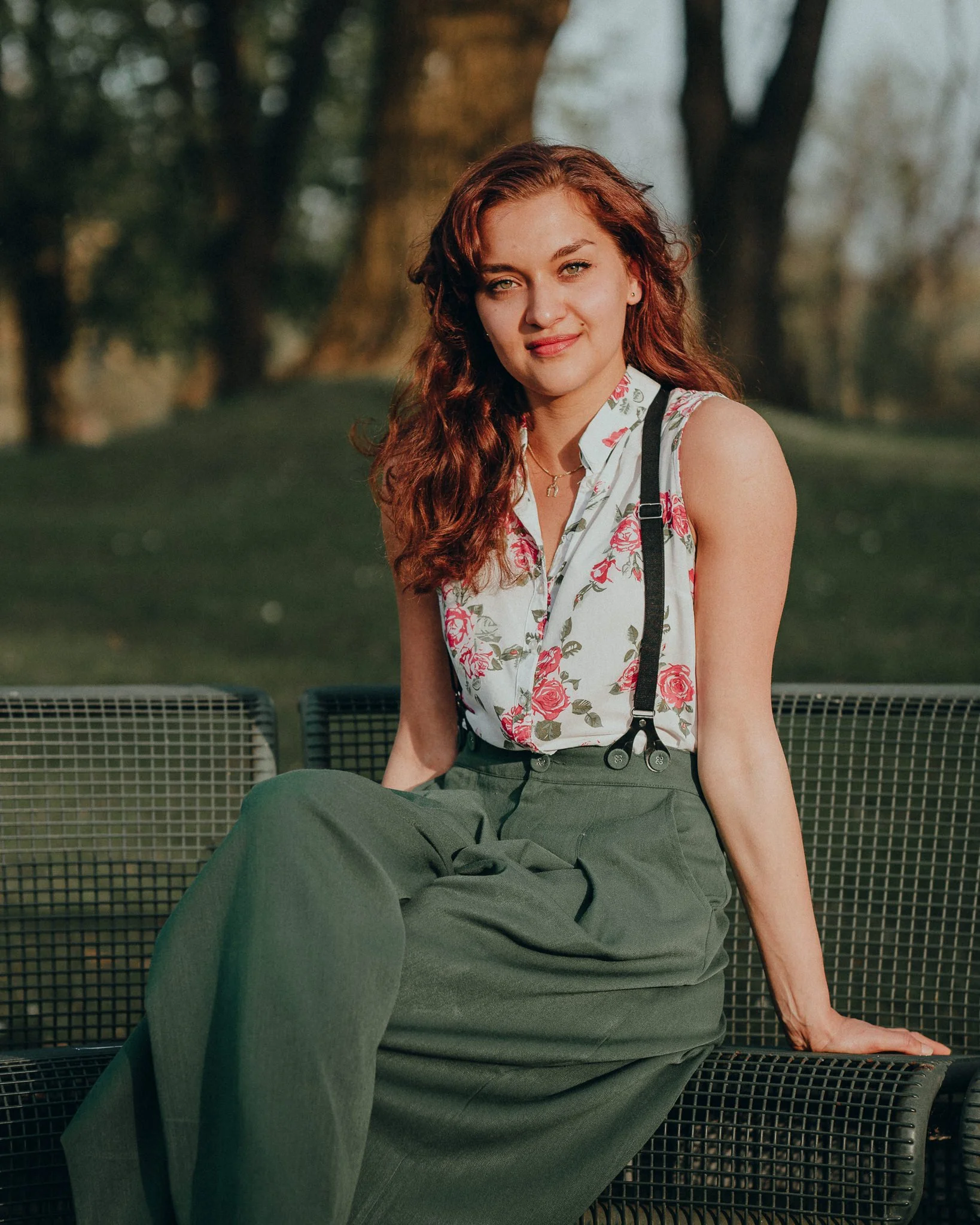 Eine junge Frau mit roten, lockigen Haaren sitzt auf einer Parkbank im Freien, umgeben von Bäumen, bei Sonnenuntergang.