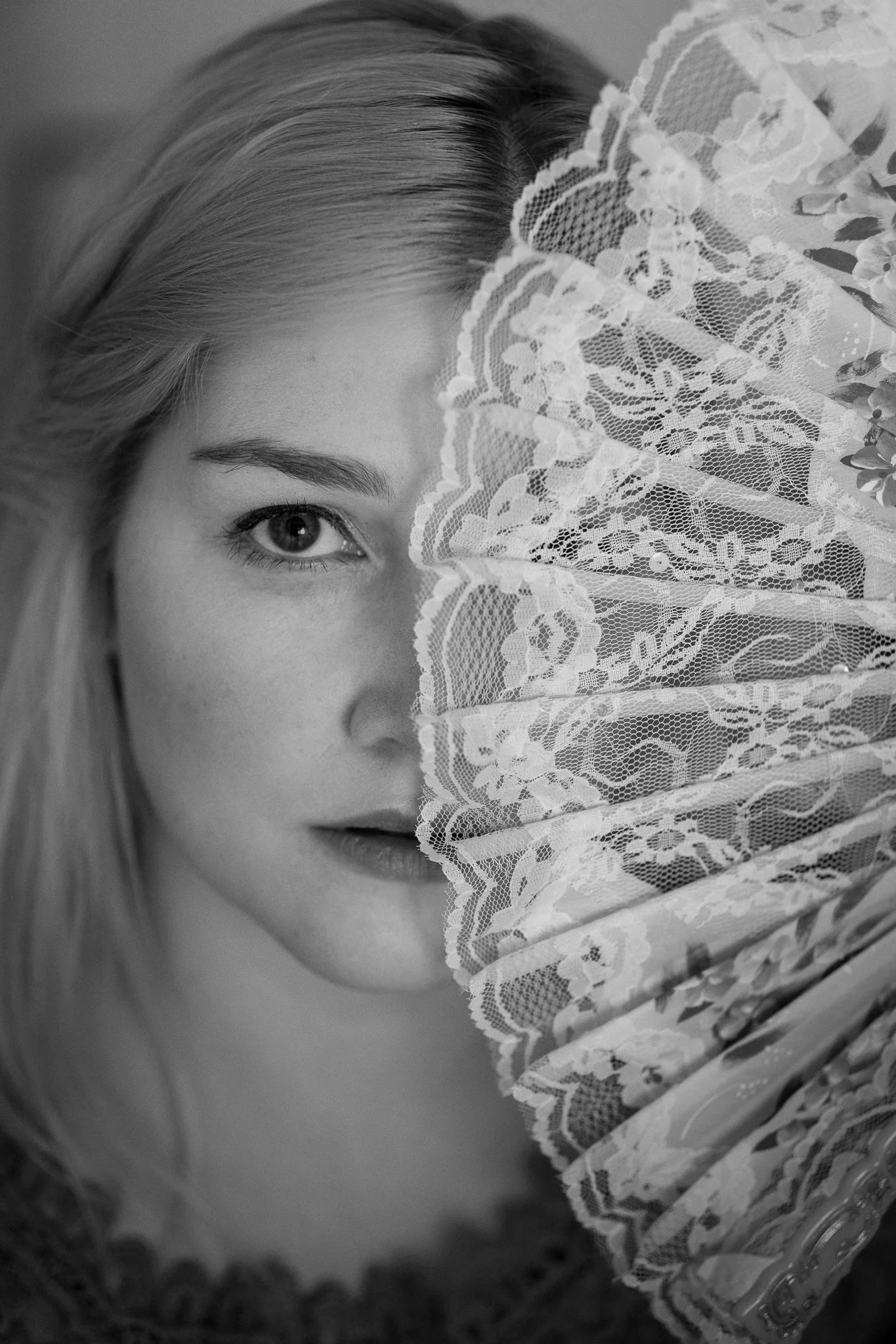 Schwarz-Weiß-Fotografie eines Mädchens mit blondem Haar, das halb von einer verzierten Spitzen-Serviette bedeckt ist, die ihr Gesicht teilweise verdeckt, nur ein Auge, die Nase und die Lippen sind sichtbar.