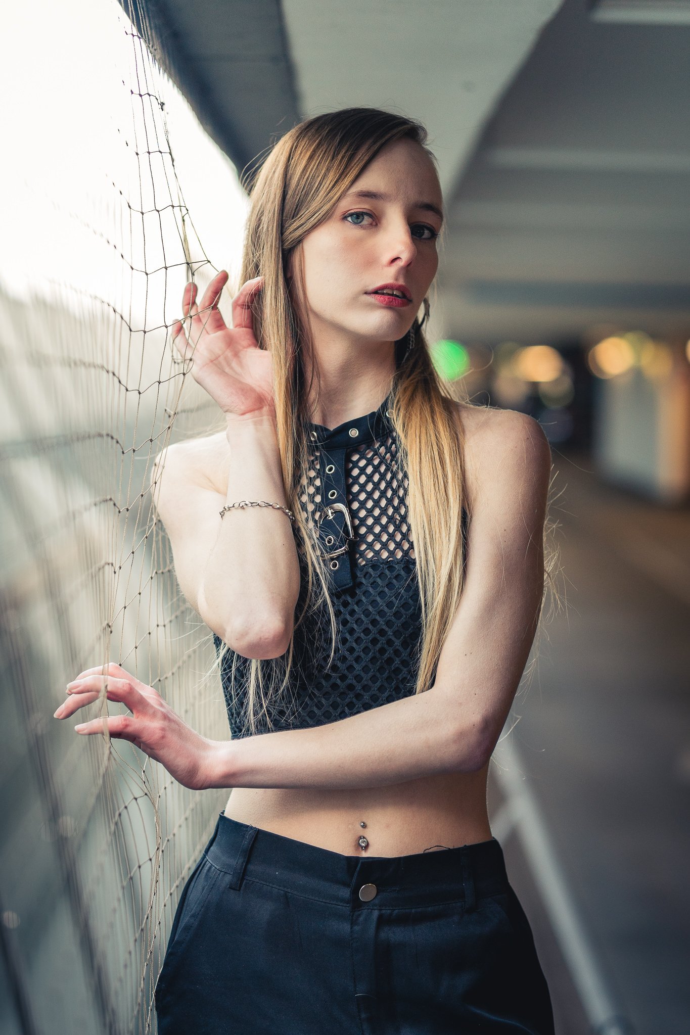 Junge Frau mit Piercing am Bauch in einer dunklen, gewerblich wirkenden Umgebung, lehnt an einem Zaun und schaut nach oben.