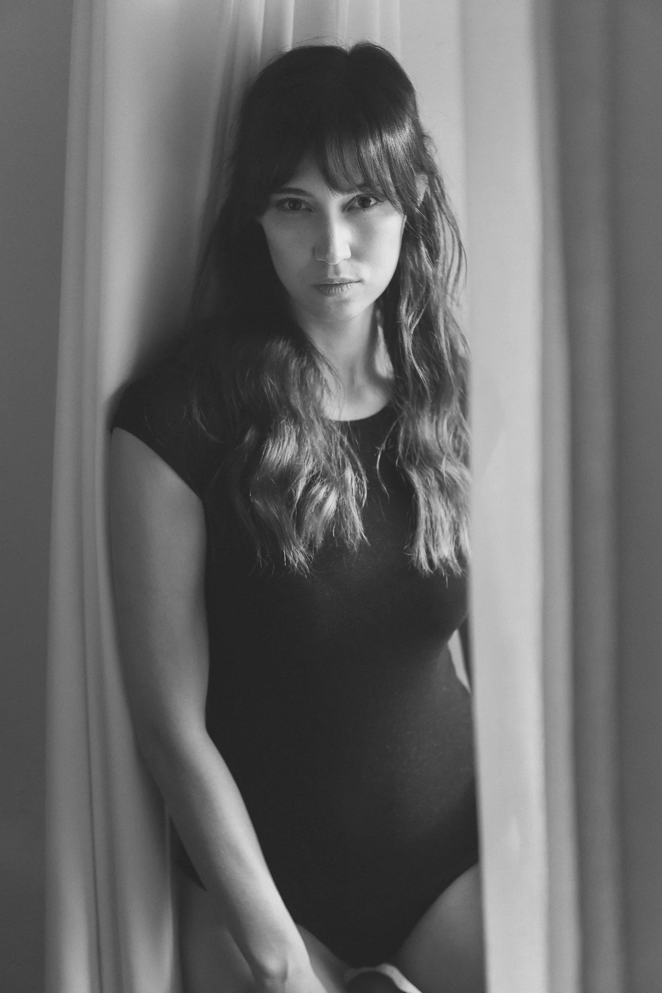 Schwarz-weiß-Foto einer jungen Frau mit langen, gewellten Haaren, die in einem schwarzen Oberteil steht und in einen Spiegel schaut.