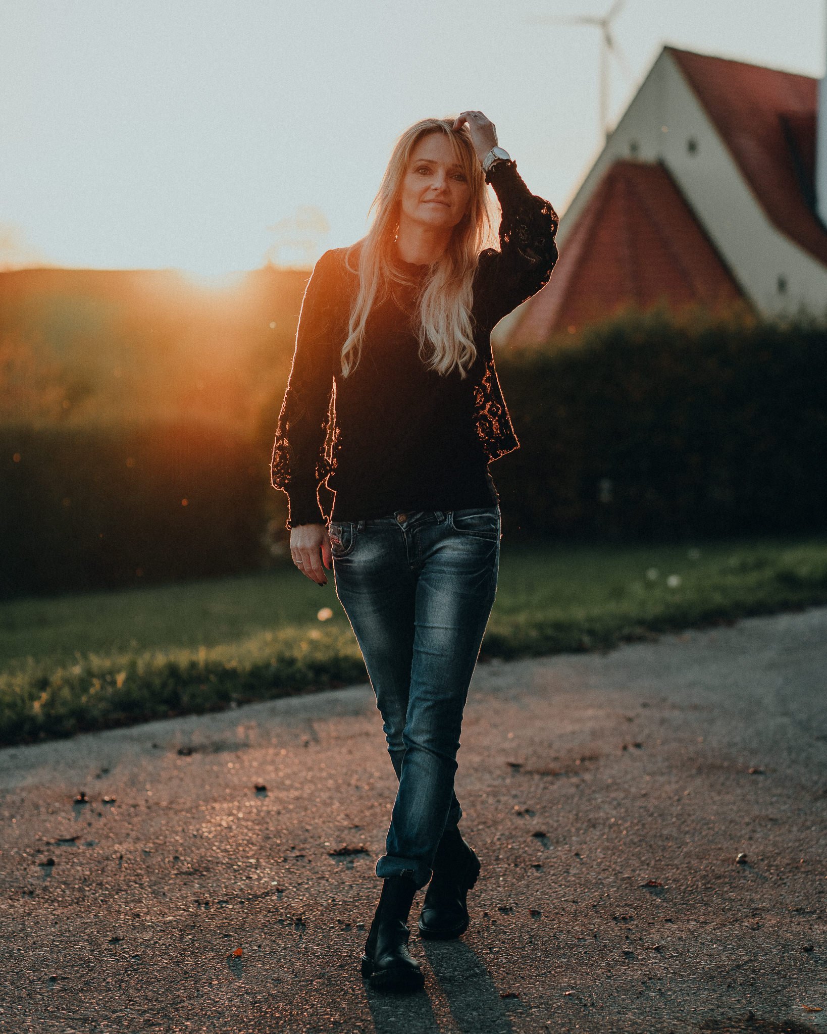 Eine blonde Frau läuft während des Sonnenuntergangs auf einem Weg, trägt eine schwarze Bluse, Jeans und Stiefel.