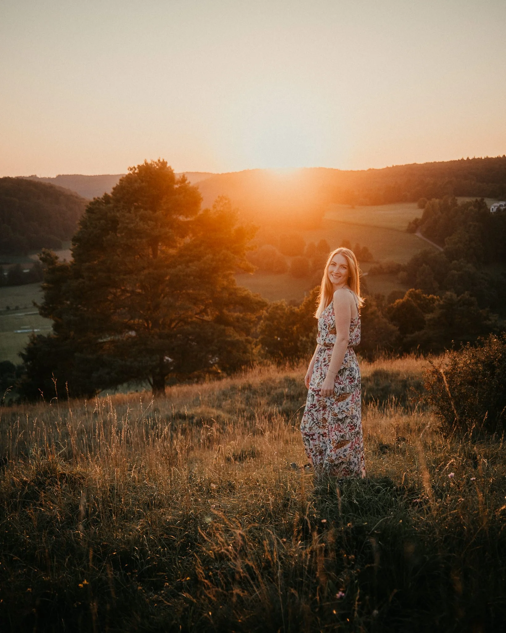 Junge Frau im Sommerkleid steht in einem goldenen Sonnenuntergang auf einer Wiese mit Hügeln und Bäumen im Hintergrund.
