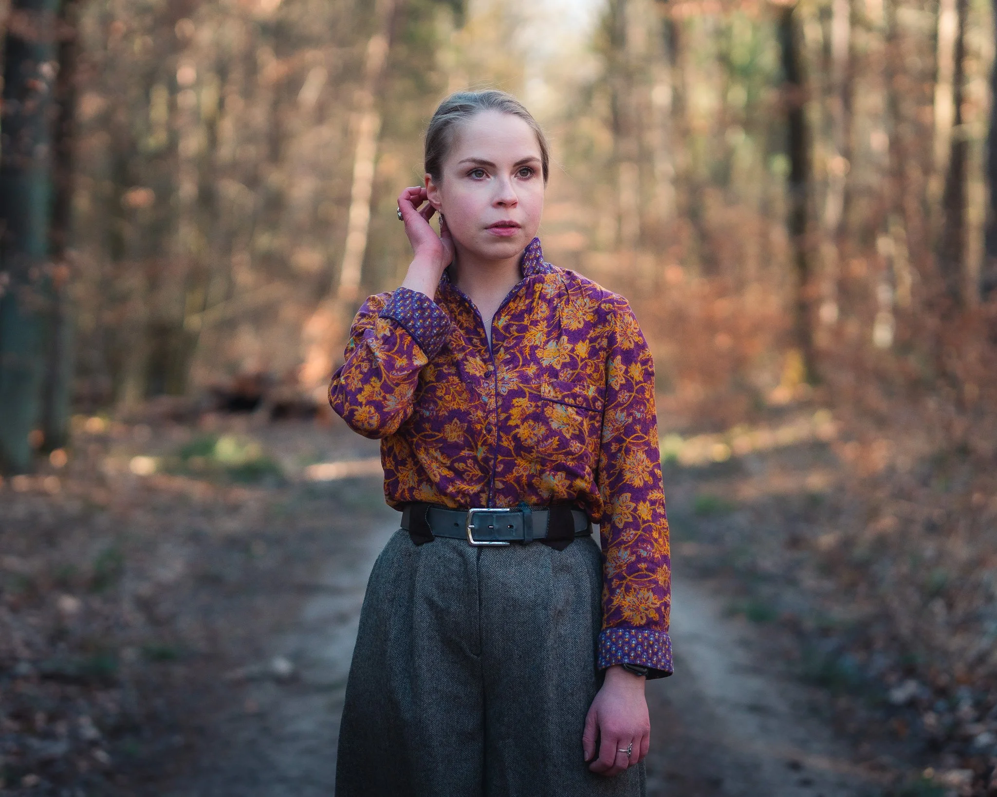 Junge Frau steht in einem herbstlichen Wald, trägt eine bunte Jacke mit Orang-, Violett- und Gelbtönen, dunkle Hosen, schaut nach rechts und berührt ihr Ohr.