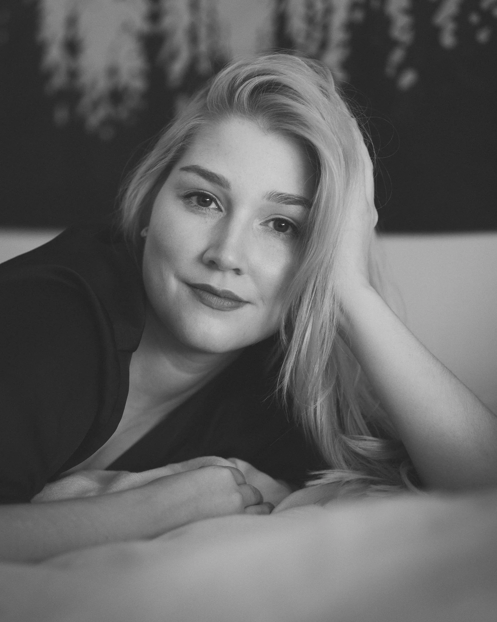 Schwarz-weiß-Fotografie einer jungen Frau, die auf einem Bett liegt, mit entspanntem Gesichtsausdruck, Kopf auf der Hand, lange Haare, die über die Schulter fallen, im Hintergrund eine Wand mit Kunstwerk.