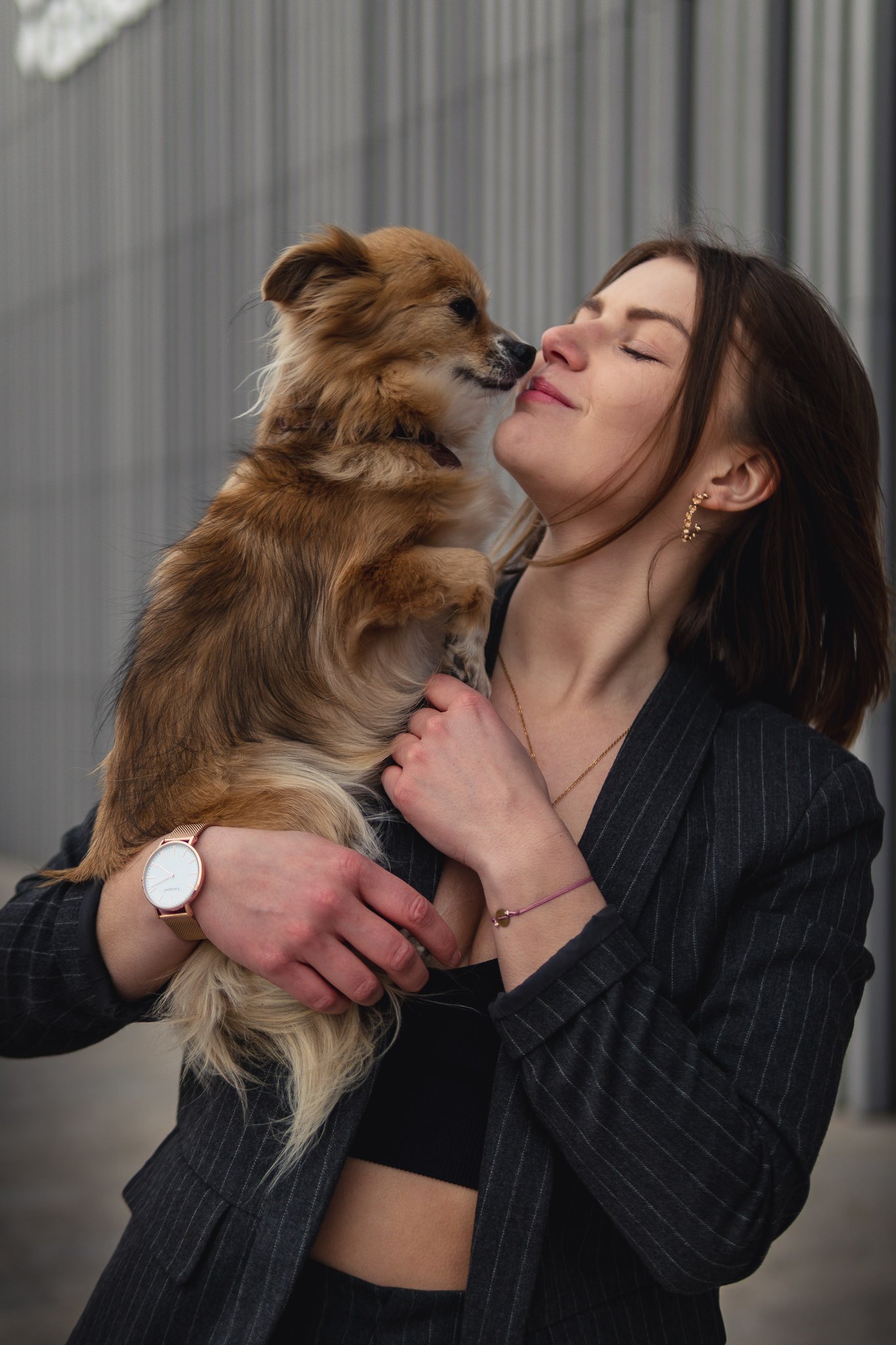 Junge Frau mit dunklen Haaren hält einen kleinen braunen Hund hoch und küsst ihn liebevoll, vor einem metallischen Hintergrund.