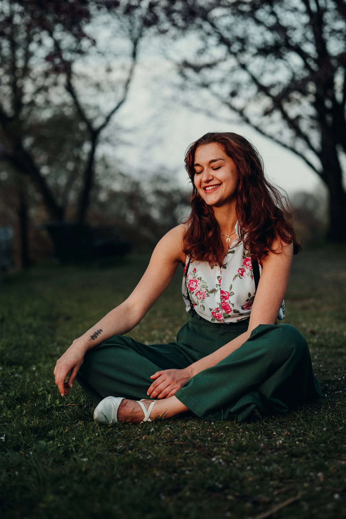 Eine lachende junge Frau mit roten, lockigen Haaren sitzt auf dem Gras in einem Park, umgeben von Bäumen, bei Sonnenuntergang, trägt eine weiße Bluse mit rosenmuster, grüne Hosen, weiße Schuhe, und hat ein Tattoo am linken Arm.