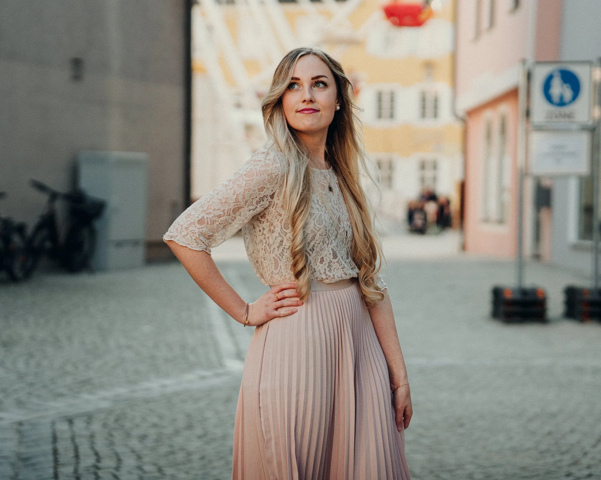 Eine junge Frau mit langen blonden Haaren in einem Innenstadtbereich mit historischen Gebäuden, trägt ein cremefarbenes Spitzenoberteil und eine hellrosa, plissierte Hose.