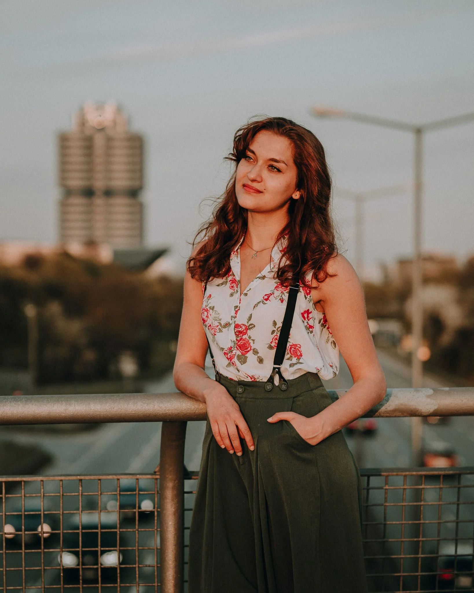 Eine junge Frau mit braunen, lockigen Haaren lehnt an einem Geländer auf einem Parkplatz im Abendlicht. Im Hintergrund sind Bürotürme und Straßenlaternen sichtbar.